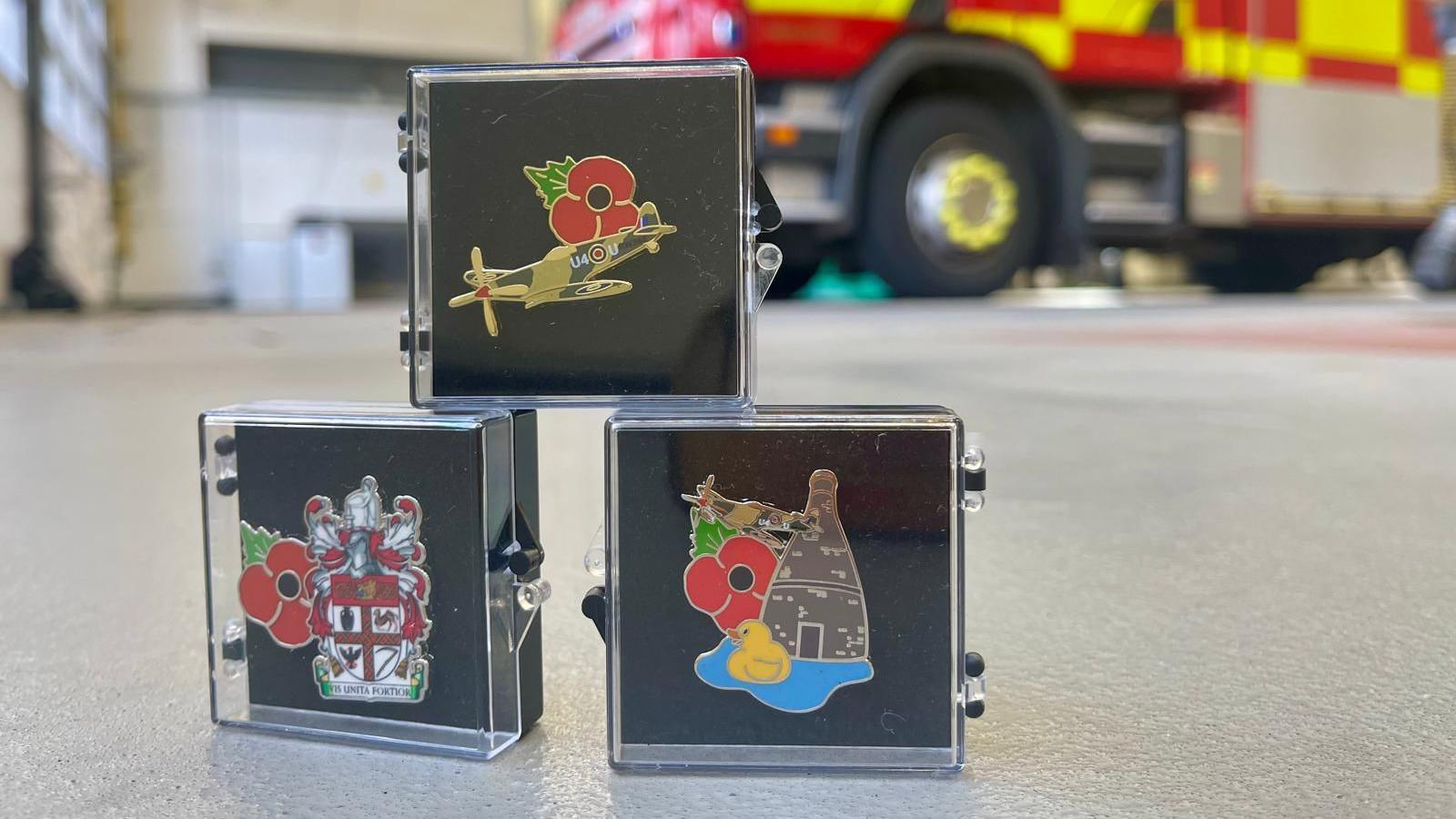 Three poppy pin badges are stacked in clear display cases. They sit on the floor of a fire station, with a fire engine visable in the background. The poppy badges include a spitfire plane, the coats of arms of Stoke-on-Trent and a combination design consisting of a spitfire, bottle kiln and a duck on a lake