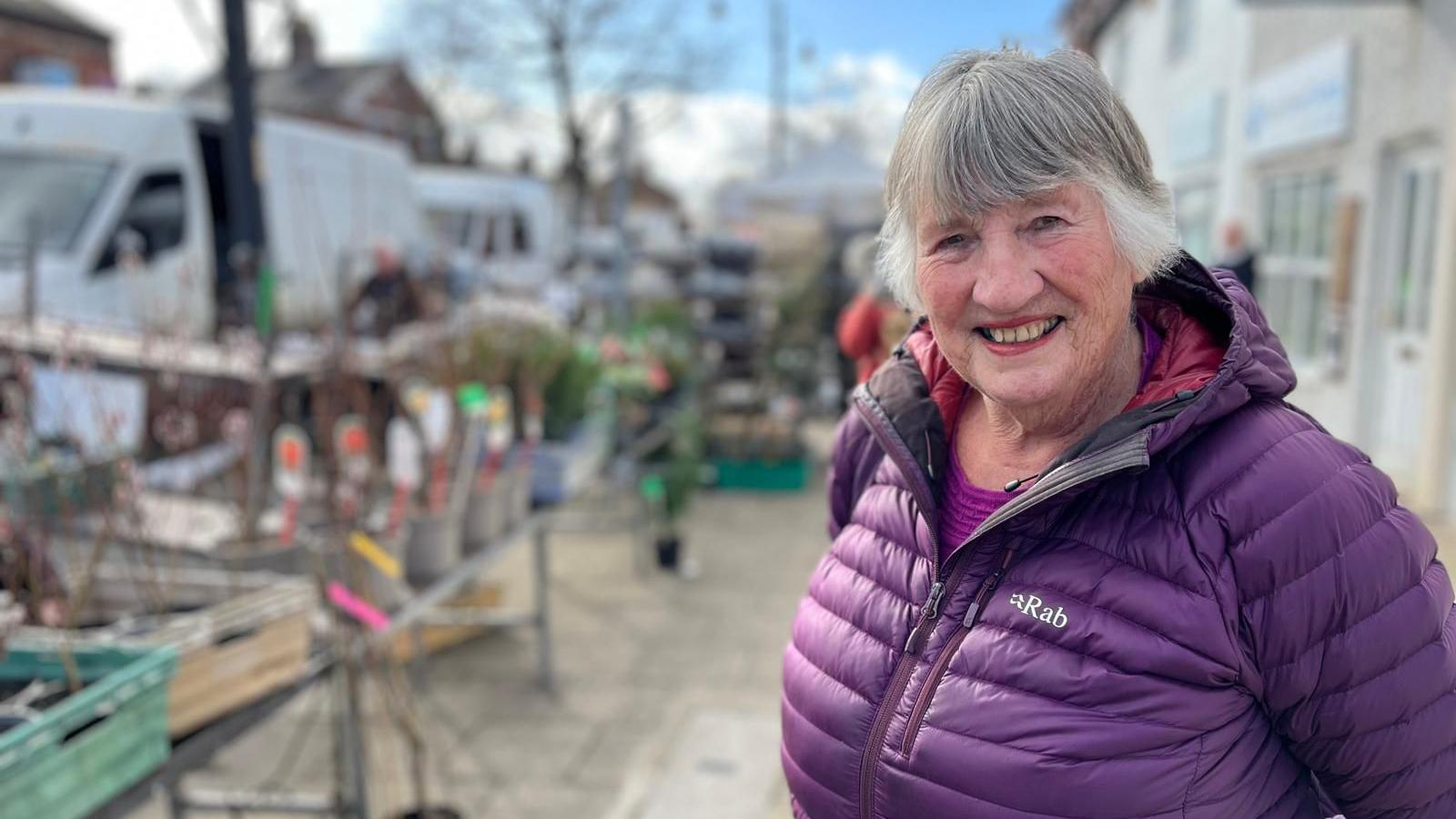 Nan Haworth, with short grey hair cut into a fringe and wearing a purple bubbl coat, smiles at the camera on a street with market stalls in the background