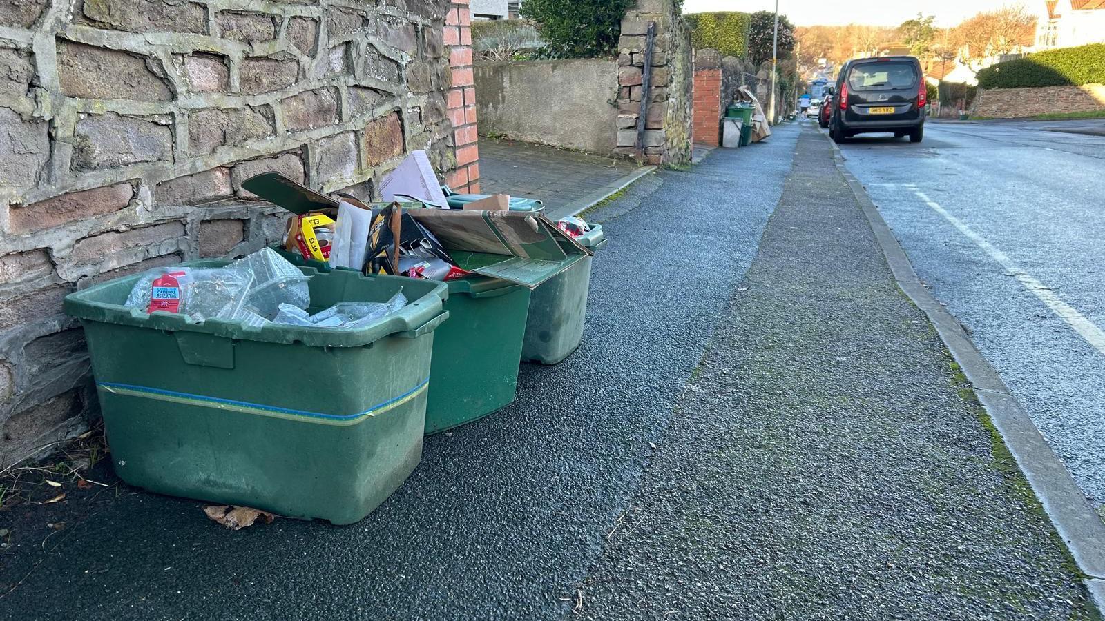 Three overflowing green recycling boxes sit on a pavement outside someone's front garden wall. They contain plastic packaging and cardboard.