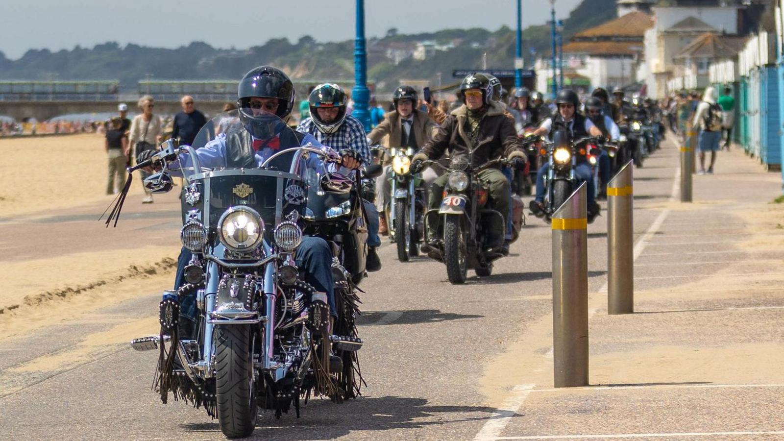 A large group of people riding motorbikes along a beachfront.