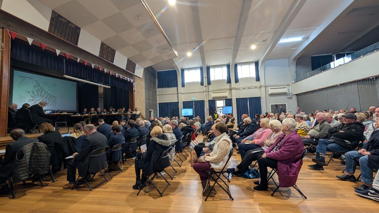 Rows of people are sitting on chairs in a hall, facing a stage which has people sitting at a table. A diagram or map is visible on a big screen.