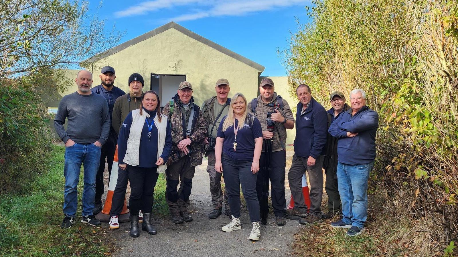 A group of volunteers, smiling, stand on the path outside the bird hide. 