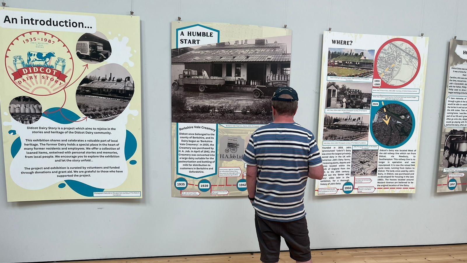 A man stands in front of a display telling the story of the dairy farm. He has his back at the camera. He is wearing a cap and a striped blue and white T-shirt and dark shorts.