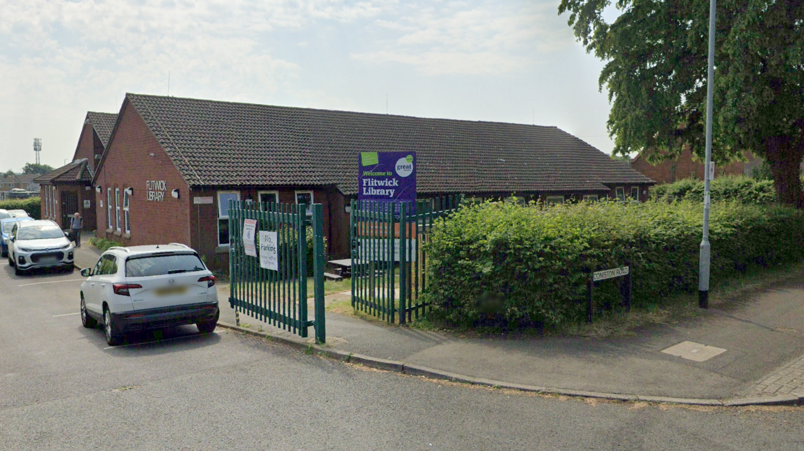 The outside of Flitwick Library. A single-storey building surrounded by parked cars on the road-side along one side and a bush and green gate on another.