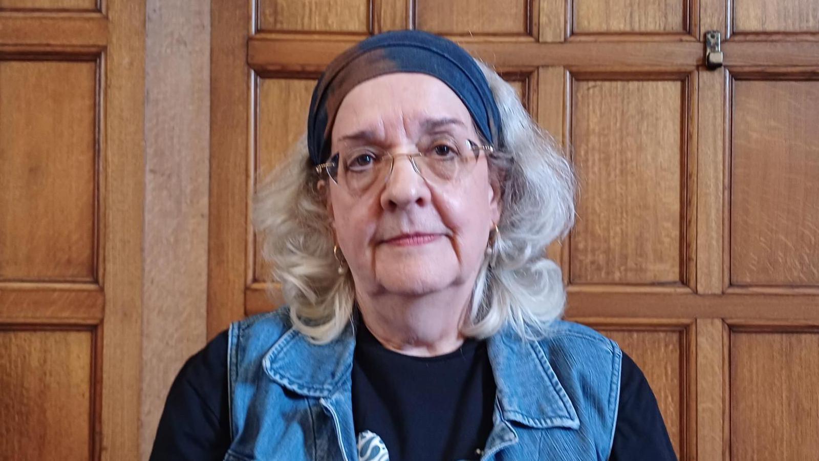 A headshot of an older woman. A polished wooden wall behind her suggests she is inside a town hall. building. She has a neutral expression and looks directly into the camera. She has grey shoulder length hair and wears glasses, hooped earrings, a navy bandana, black top and denim waistcoat.