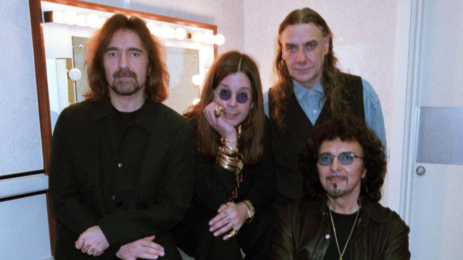 Butler, Osbourne, Ward and Iommi, dressed in black, sitting around a lit dressing room mirror. Their expressions are solemn as they look at the camera.