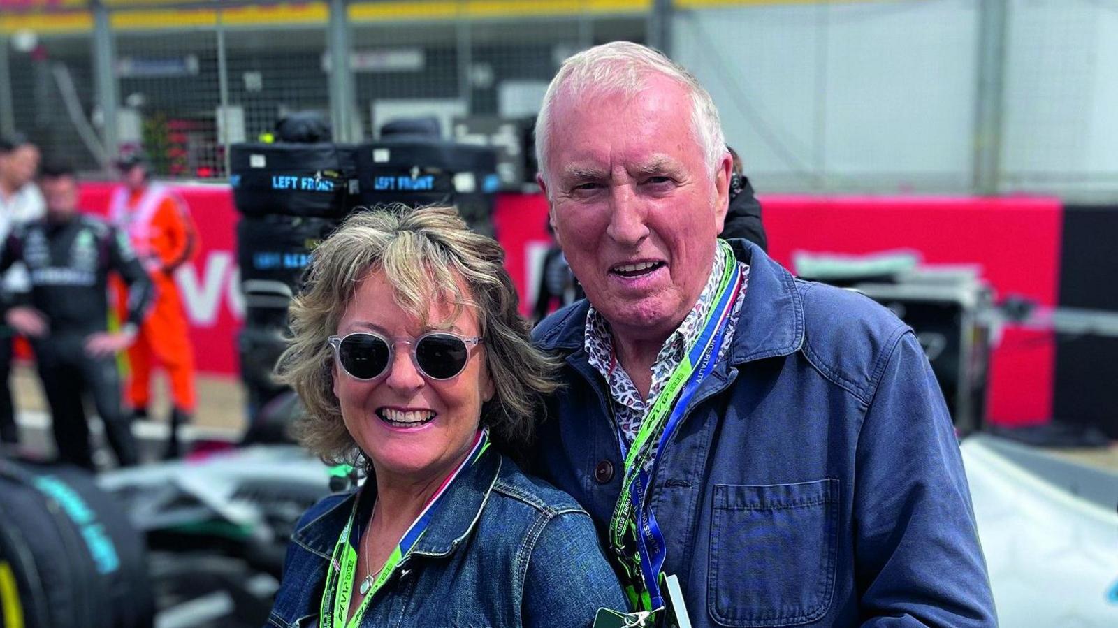 Tiggy Walker, wearing sunglasses and smiling in a denim jacket stands closely with Johnnie Walker - he also wear a denim jacket. They both have lanyards around their necks - they are at a racing circuit and you can just see formula one cars in the background. 