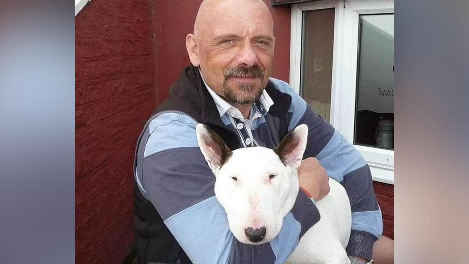 A man with a bald head and goatee beard looking at the camera and holding a white dog with black ears.