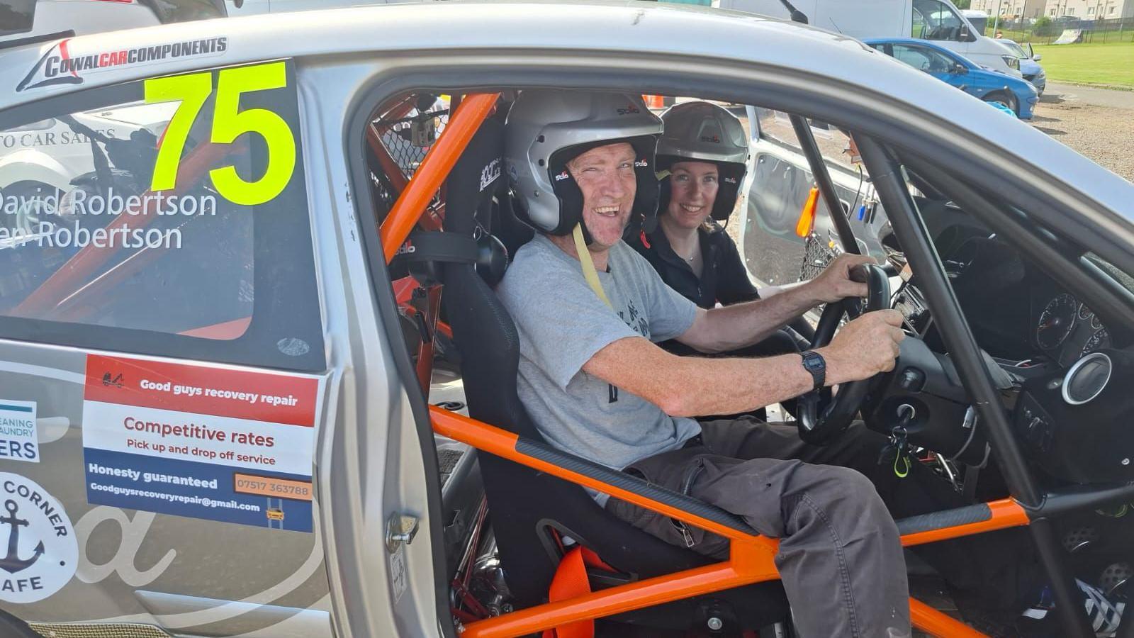 a man smiling wearing a helmet sitting inside a rally car, next to him is a woman who is also wearing a helmet.