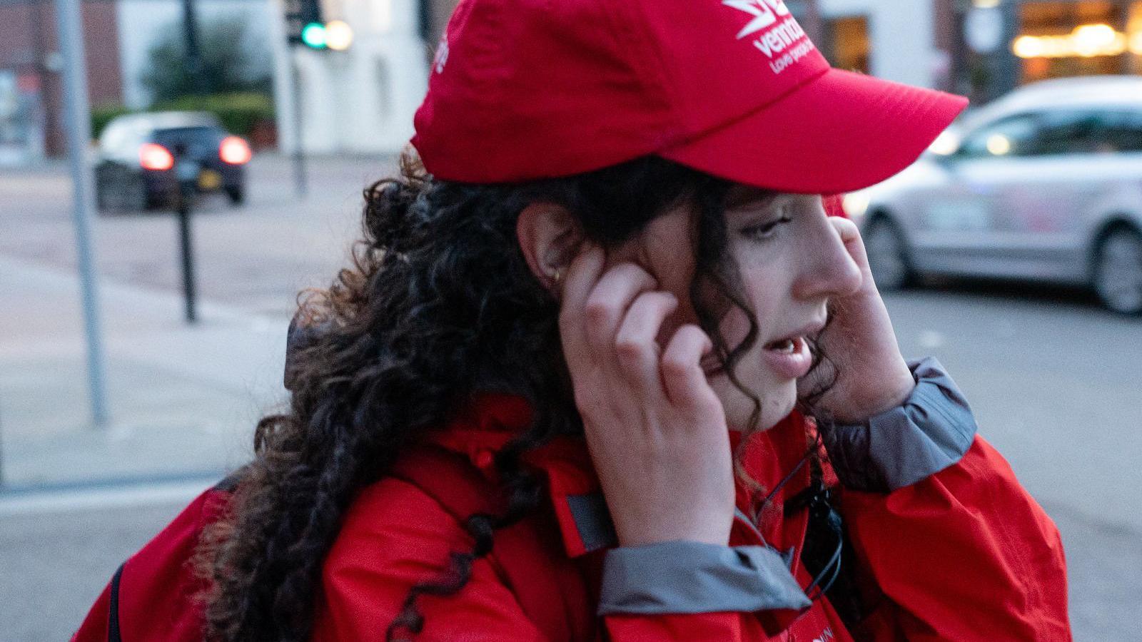 A woman with long curly brown hair wearing a red jacket and red cap. She has her hand in one ear as she speaks into a mobile phone