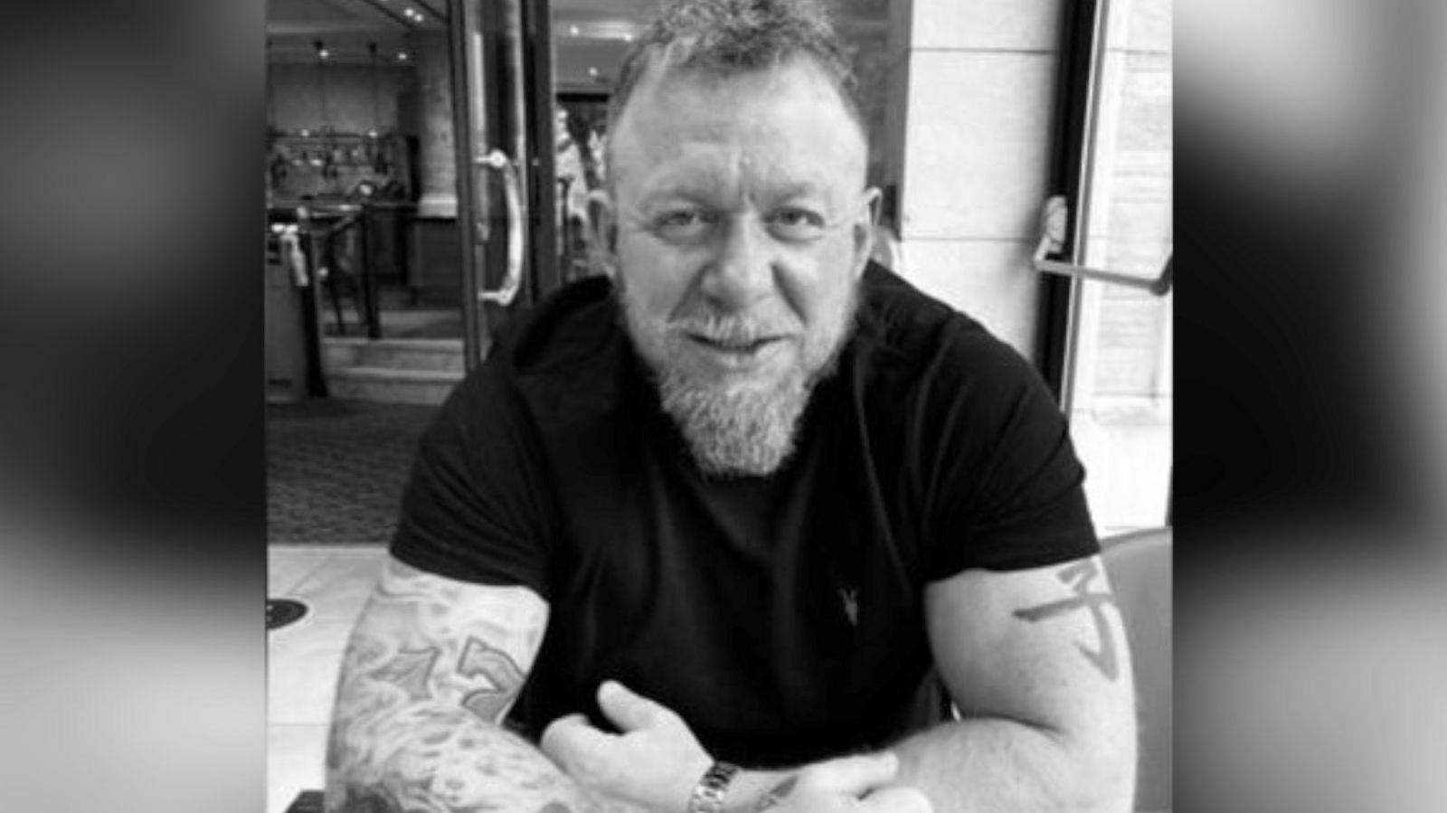 A black and white photo of Killick, who is shown in what appears to be a bar or restaurant. He is wearing a black T-shirt, showing various tattoos on his arms, and is smiling. He has a beard with longer hair at his chin.
