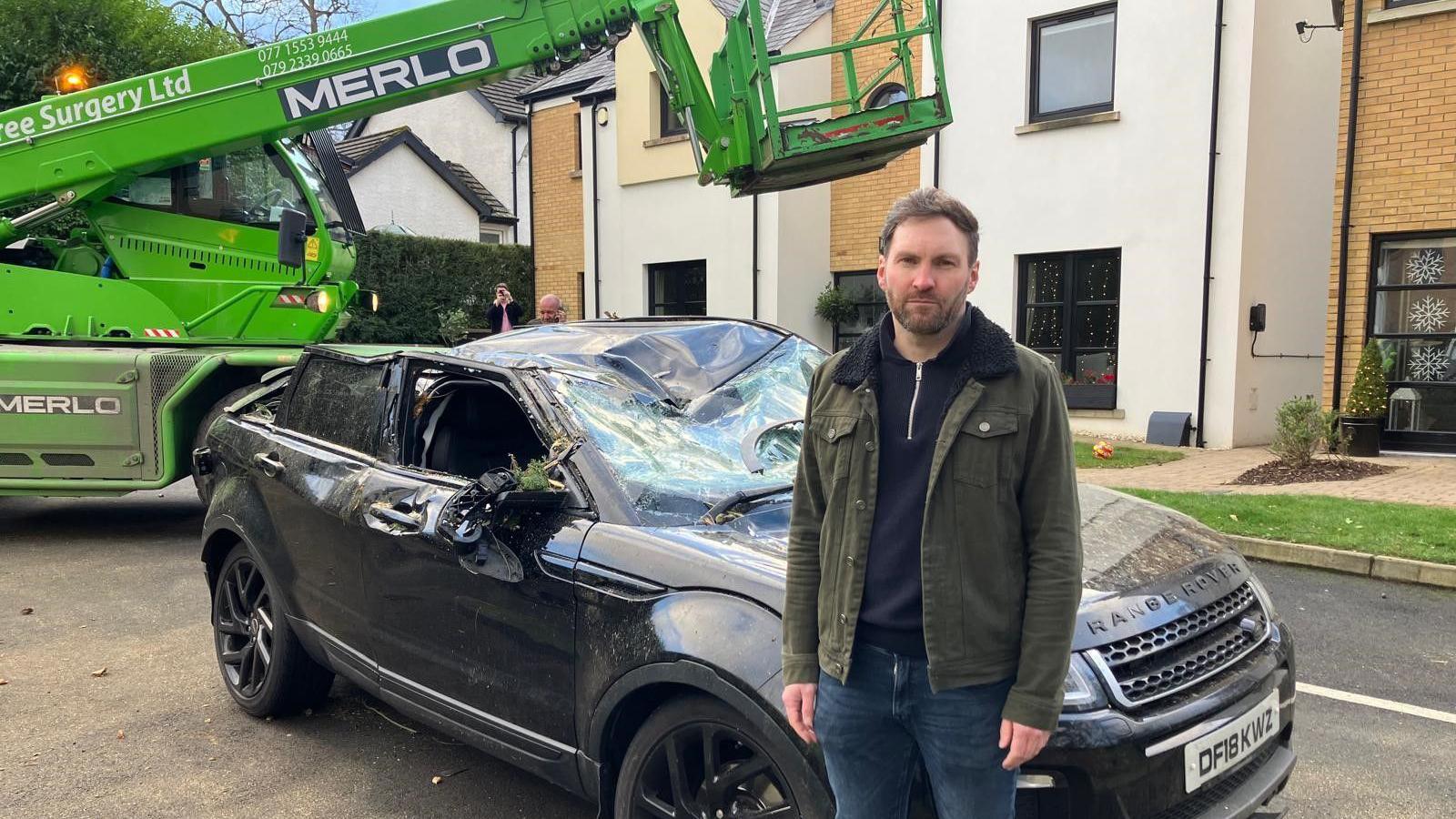 Stephen Haughey in blue jeans, a navy half-zip and a green denim jacket, stood in front of a badly smashed black range rover, the windscreen and roof have caved in, a bright green cherry picker is parked behind it... there are several new-build style houses with cream exterior walls, tan brickwork, a paved walkway and patches of greenery. 