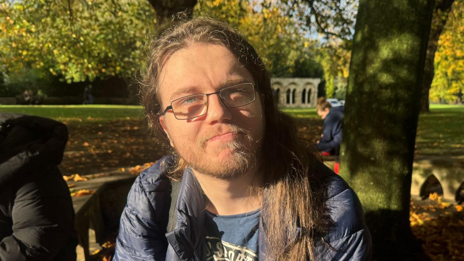 A man with long brown hair and glasses sits on a wooden bench in a park. He wears a blue jacket over a graphic T-shirt and looks toward the camera. Behind him, a large tree with autumn-coloured leaves casts shadows across the grassy area.