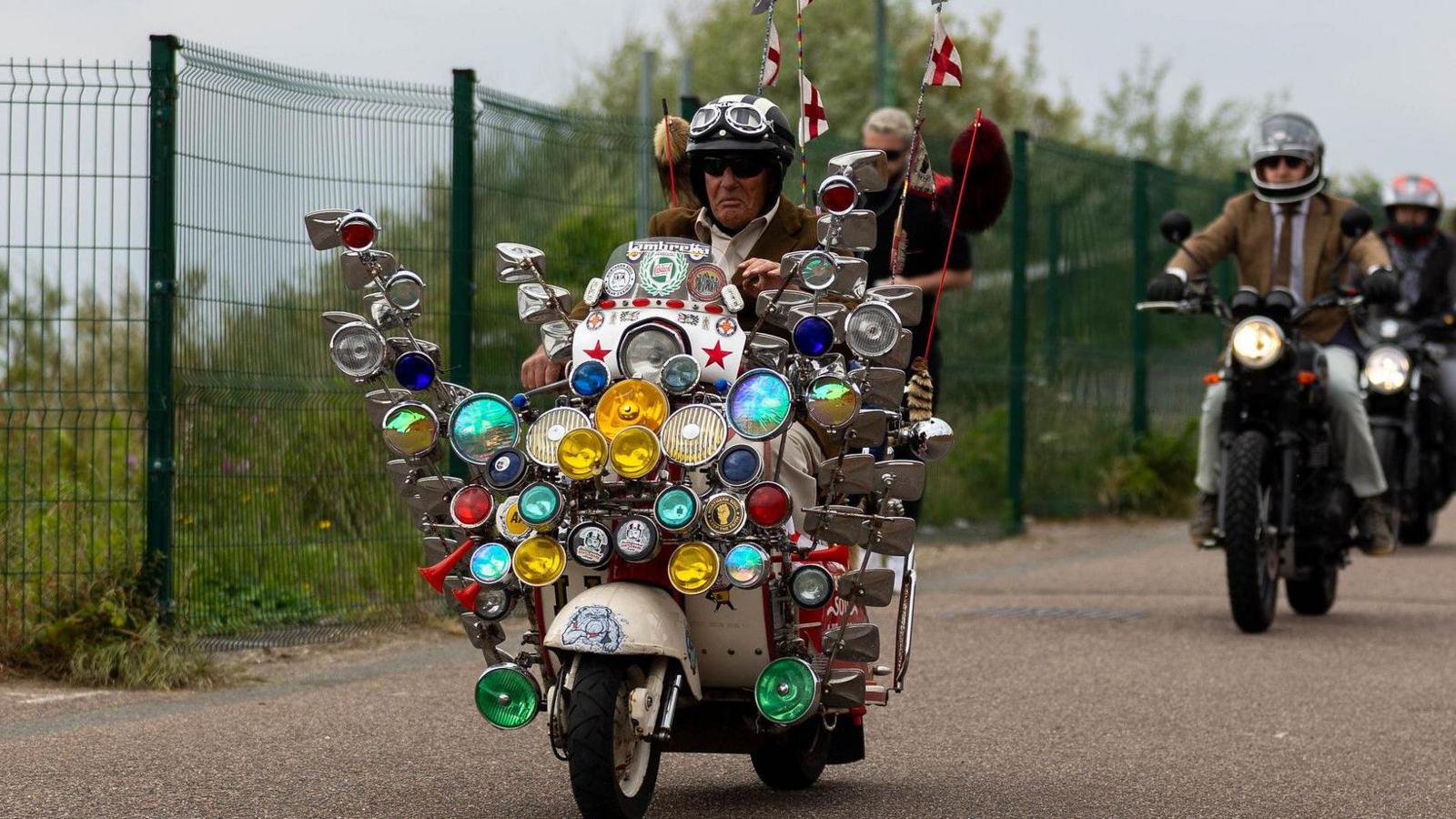 A man riding a scooter with loads of lights and mirrors at the front - like something from Quadrophenia.