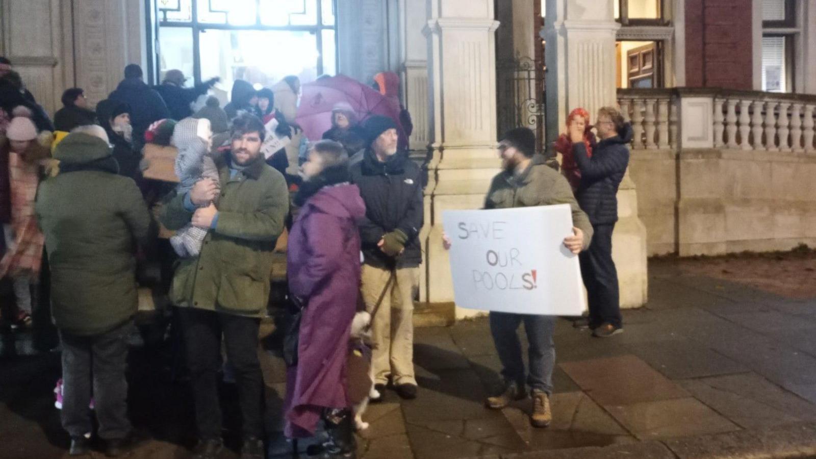 A group of protesters gathered outside. One is holding a sign which says 'save our pools'.