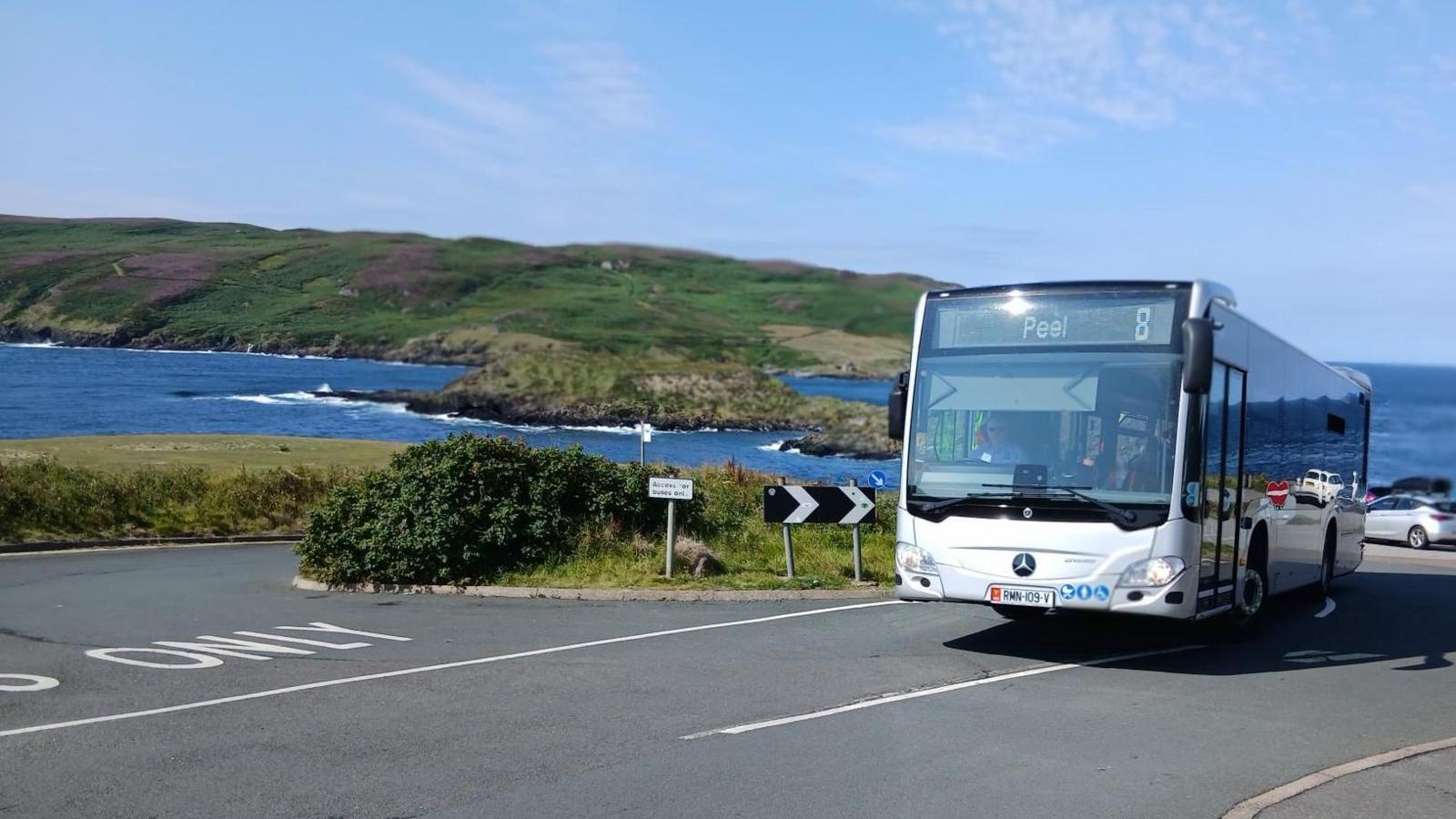 A silver bus bearing a sign reading Peel, and the route number 8. The sea and the Calf of Man can be seen in the background.