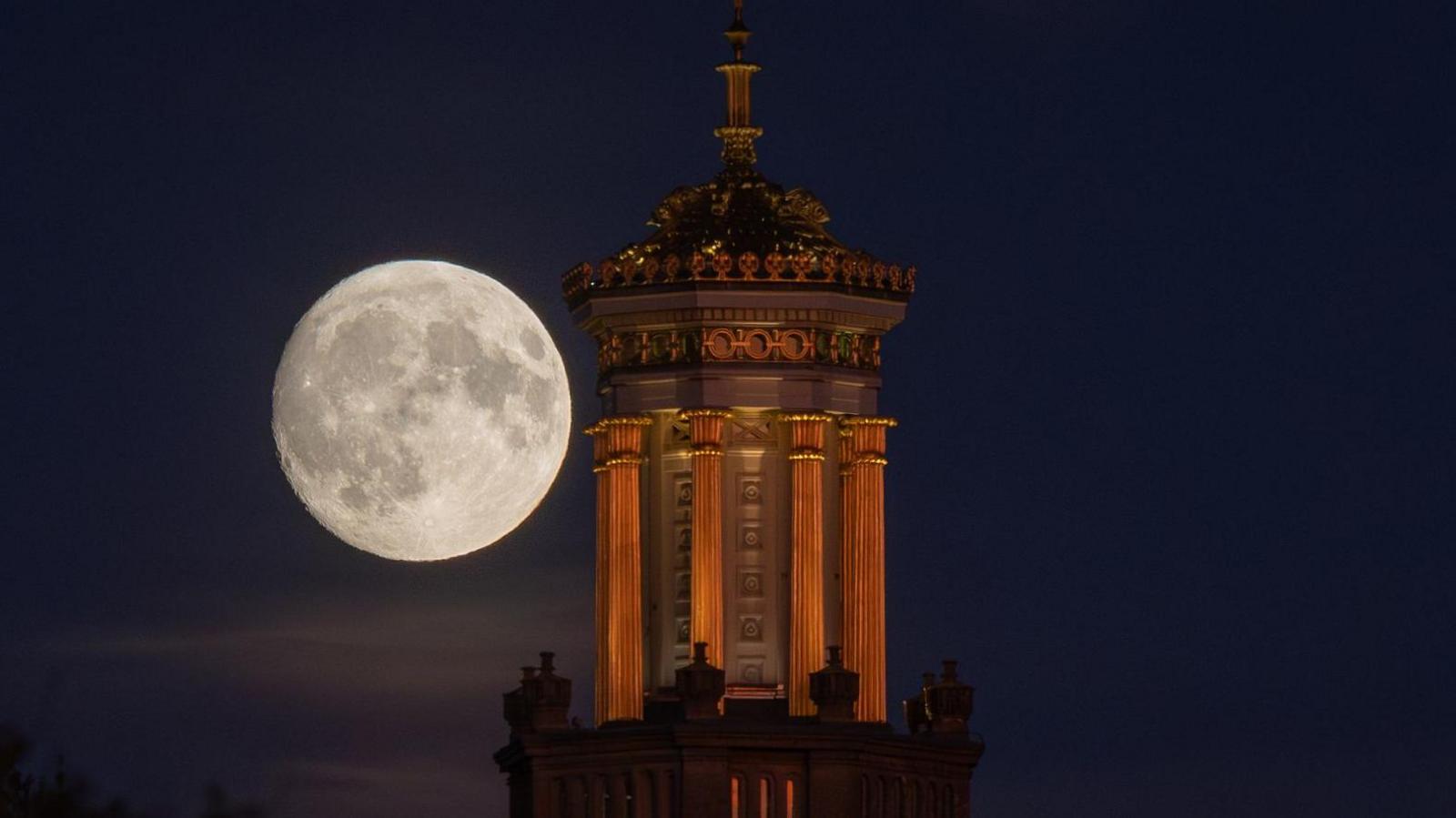 The large bright supermoon is seen behind the Beckford's Tower in Bath at night. The tower is illuminated by floodlights and the landmarks on the surface of the moon are visible