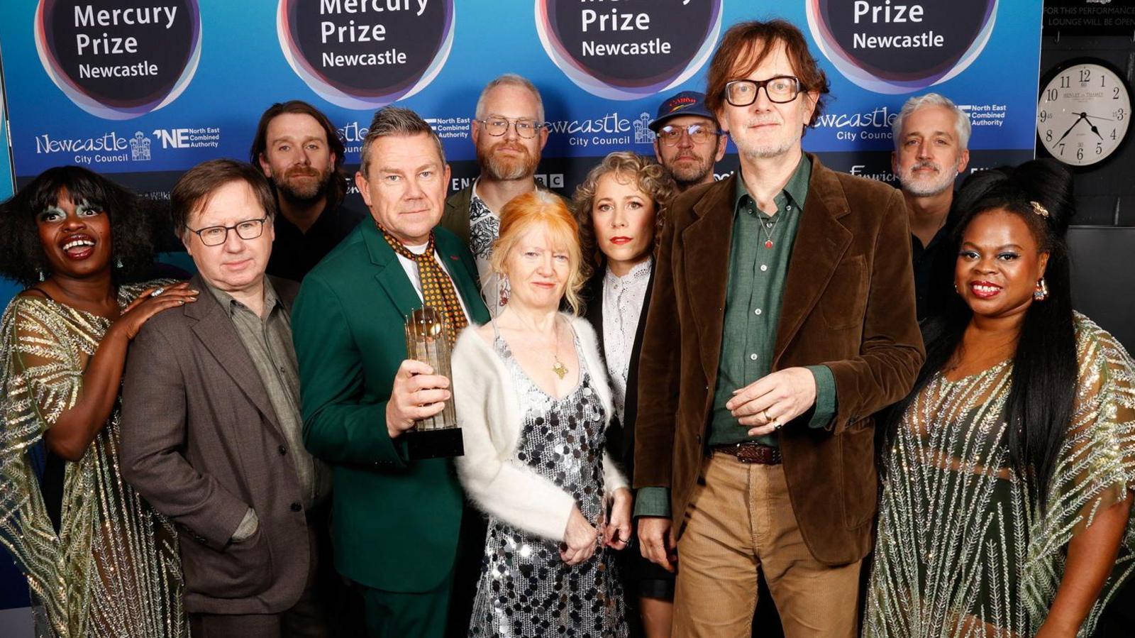 The band Pulp and accompanying musicians smiling into the camera. There are 11 people dressed in colourful clothing. Jarvis Cocker is standing at the front, second from the right and has brown, sweepy hair and is wearing large glasses. He is wearing a brown cord blazer and a green shirt underneath. He is wearing lighter brown trousers. They are standing in front of a blue Mercury Prize board.