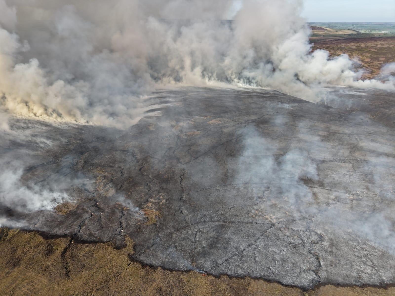 A drone shot taken above the wildfire, showing a large area of black ground and smoke.