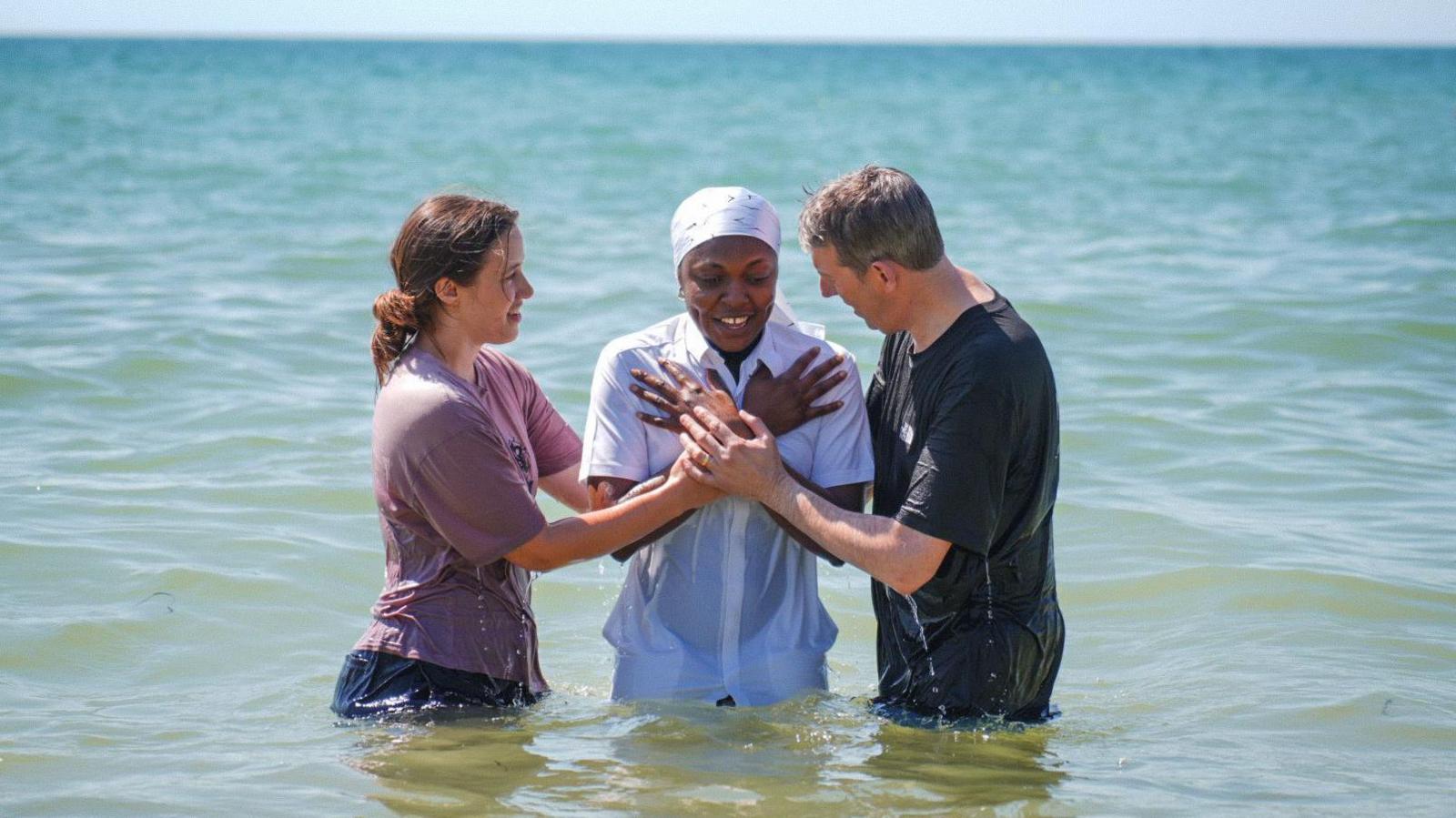 Three people in the sea two either side of a person in all white who has been dunked in the water
