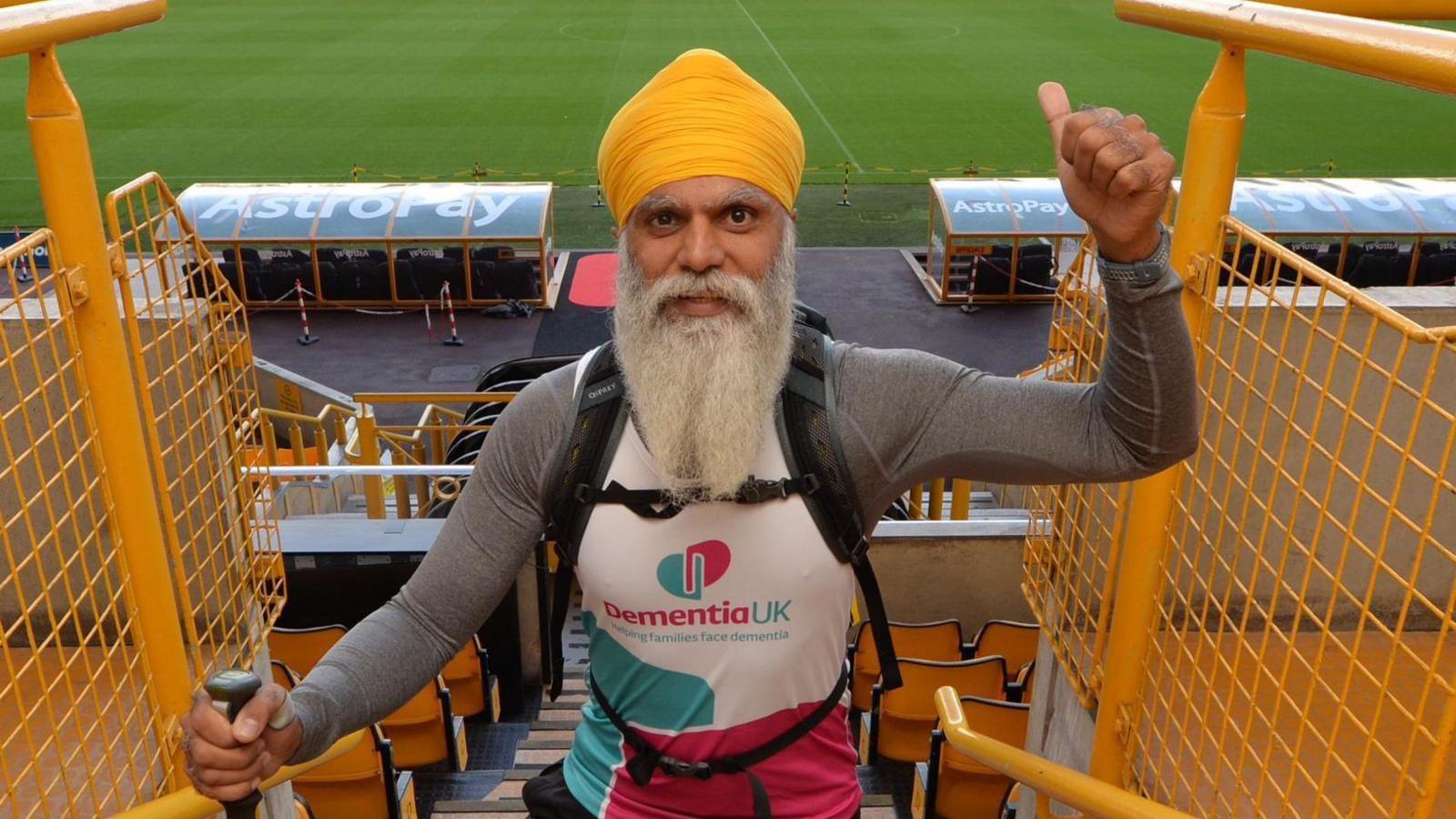 Manny Singh Kang has a long grey beard and orange turban. He is wearing a shirt that reads Dementia UK and has his thumb up. He is standing on the steps of a football stadium.