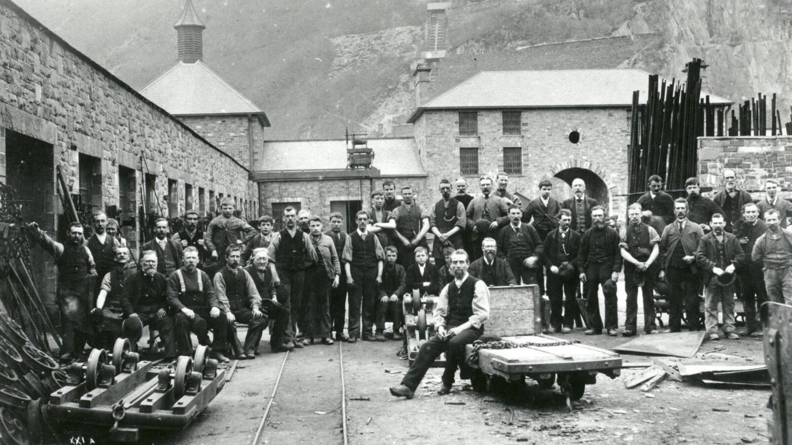 Old black and white photograph of the quarry workshops with dozens of staff posing in the courtyard amid slate equipment