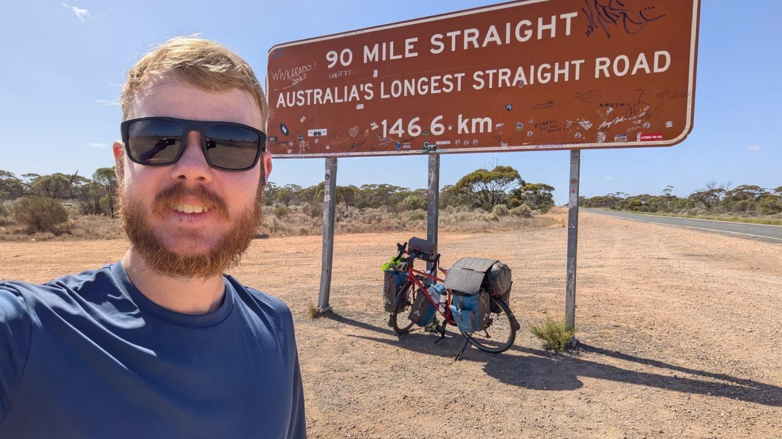 A red-haired man wearing sunglasses smiles at the camera and is stood in a dessert, in front of a road sign which reads 'Australia's longest straight road'. His bike is parked in front of the sign.