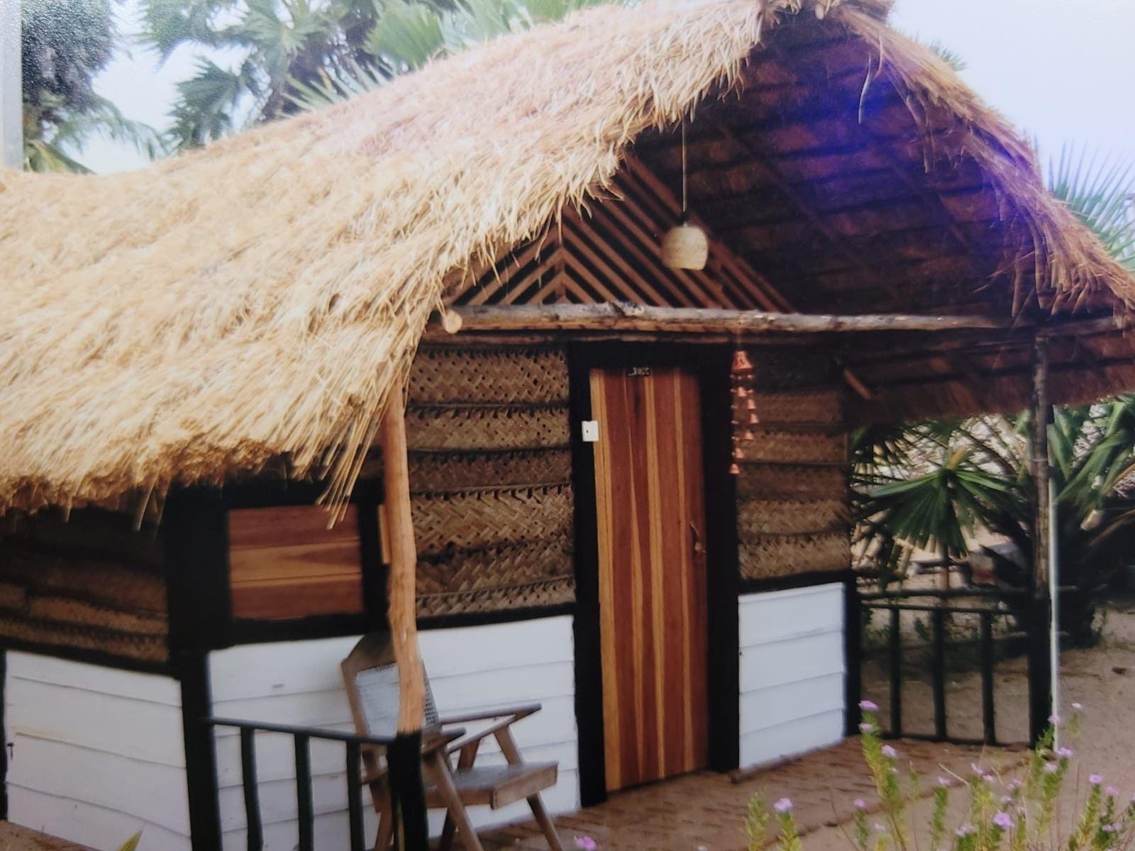 A small thatched-roof wooden hut with woven walls and a wooden door sits on sandy ground, surrounded by tropical plants and trees.