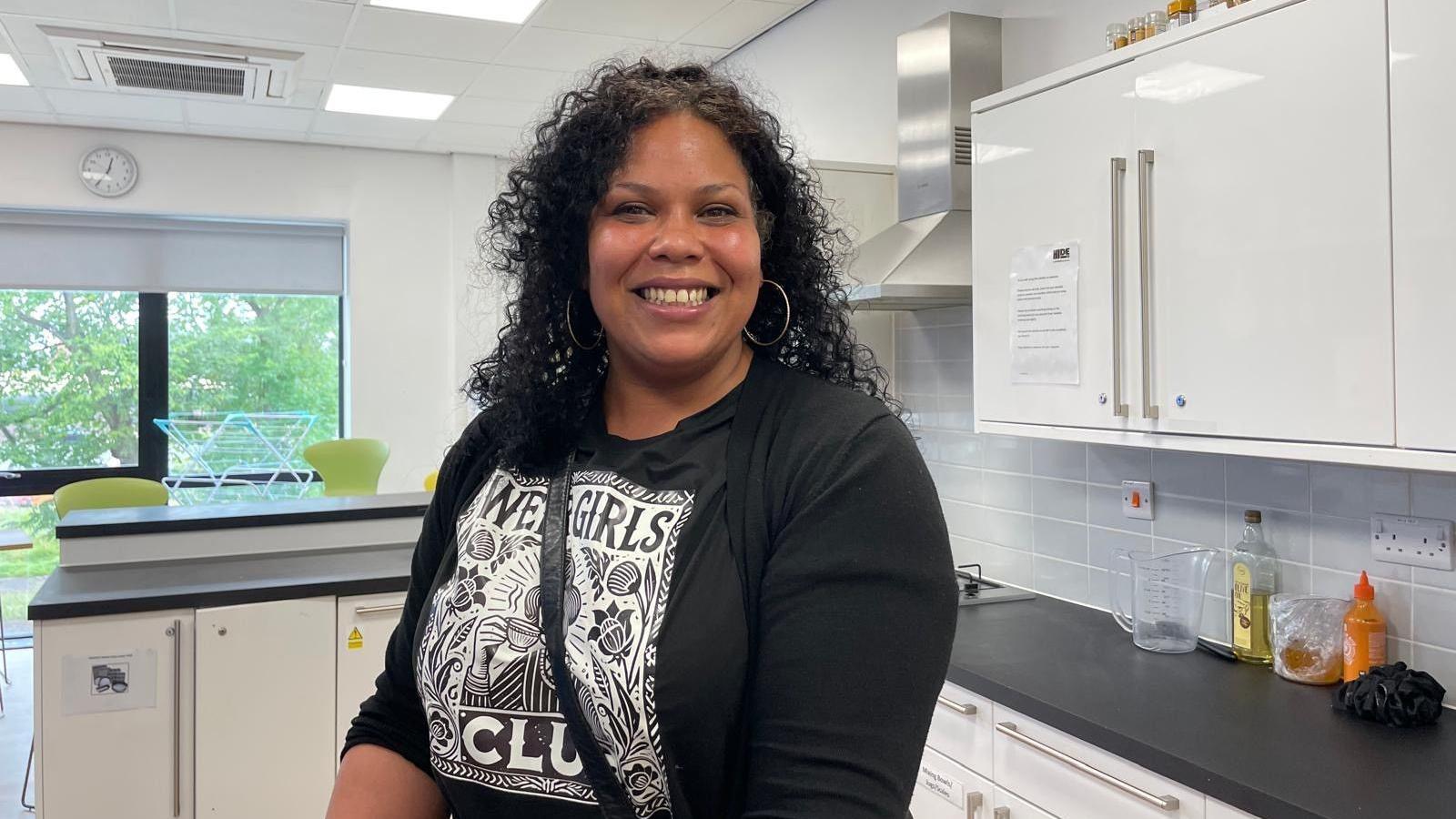 Evelyn with shoulder length black hair in tight curls wearing a long sleeve black top with white floral design in a big square on the front. She is standing in a community kitchen smiling at the camera.