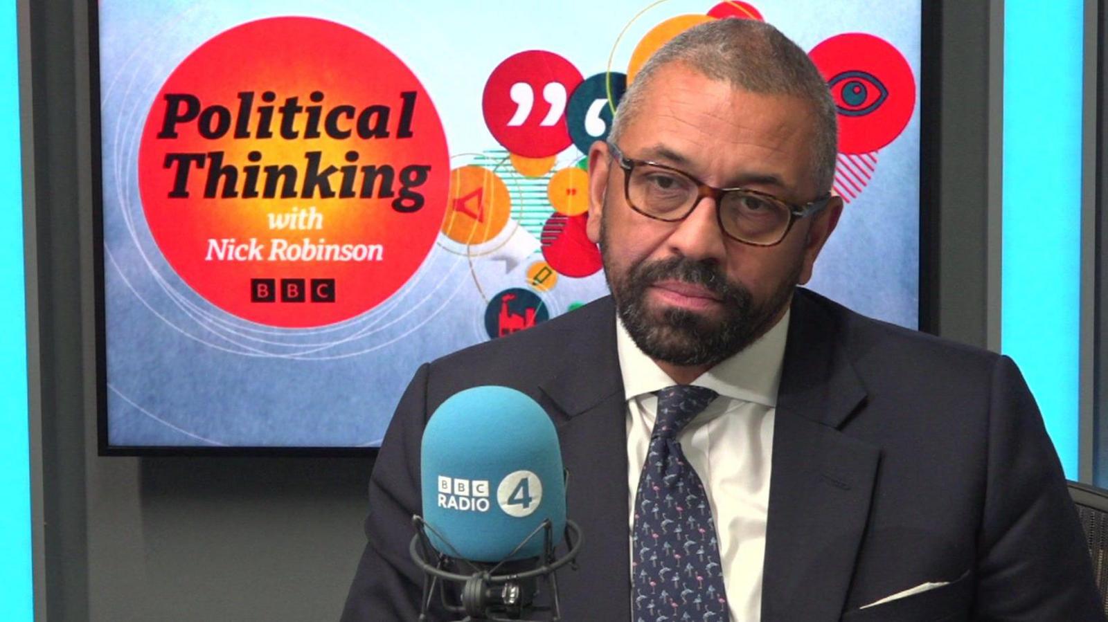 James Cleverly wearing a suit sits in front of a sign for Political Thinking with Nick Robinson while recording his interview.