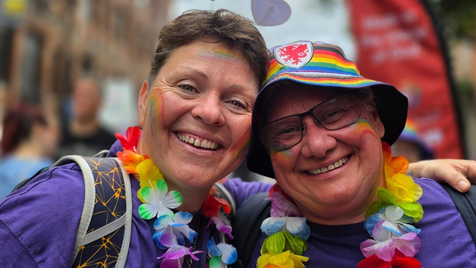 Two women with rainbows painted on their cheeks, wearing purple tops and floral necklaces smiling. The background is blurred but it appears they are at a parade for an LGBT+ event.