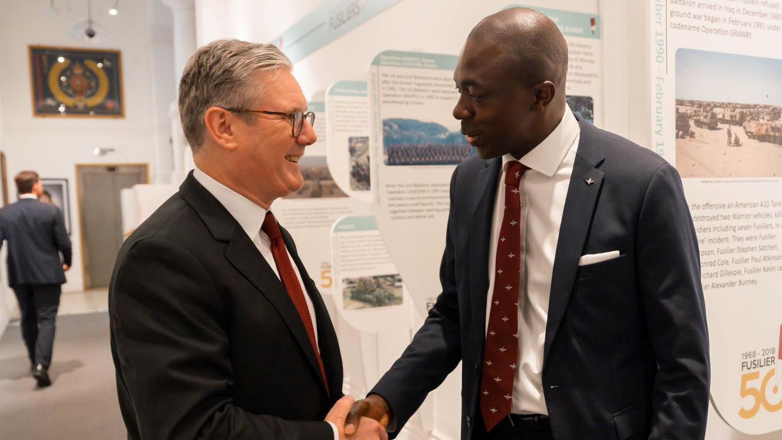 Two men shaking hands. Both are wearing navy jackets with a deep red-coloured tie. They are at an event to celebrate the military and there are boards with photographs behind them in the near background.