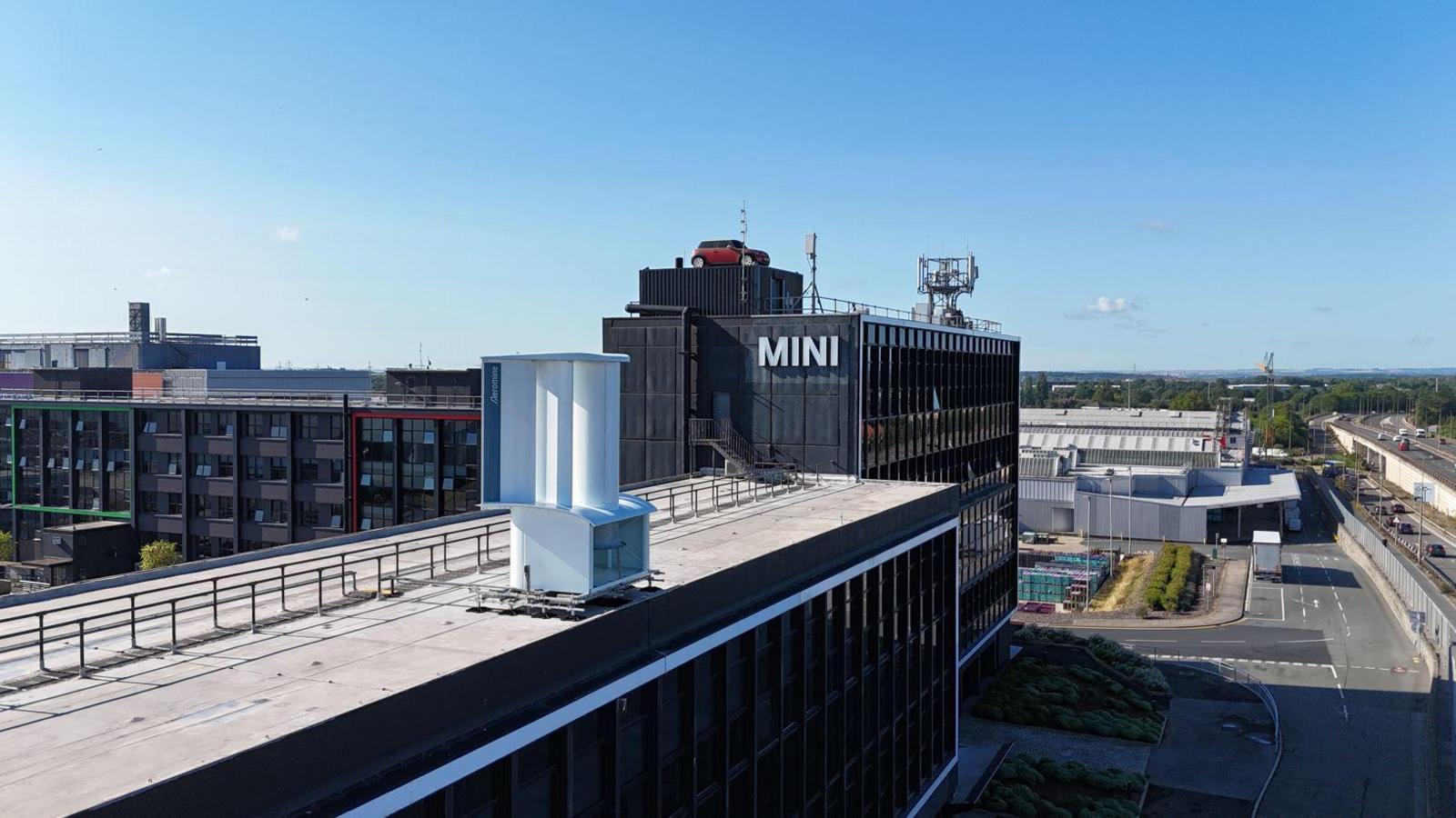 The Mini Plant in Oxford photographed from a bird's eye view. The new wind  unit is also seen on the edge of a nearby building. The name of the Mini plant is seen, as well as a red car on top of the building. It's a clear day.