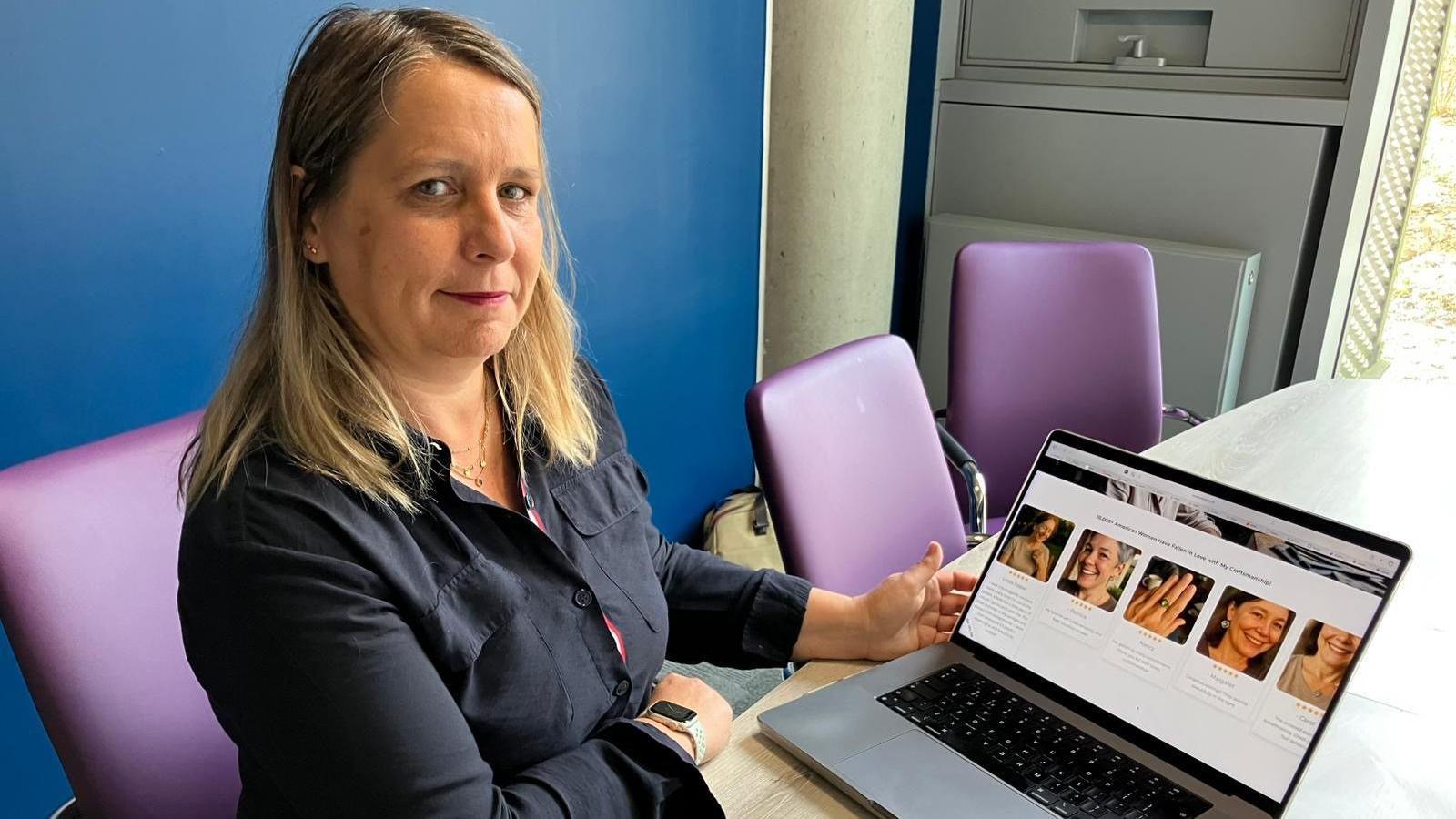 A woman in a dark shirt sat at a table on a purple chair. On the table is an open silver laptop displaying images from the Scarlett Jewels website, likely generated by AI.