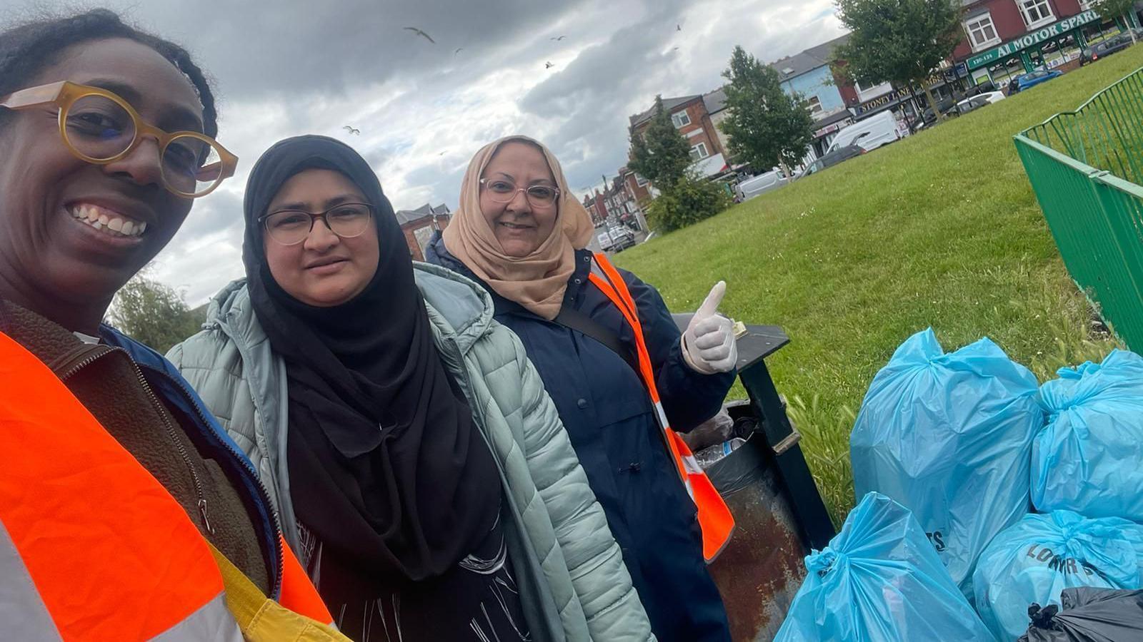 Three women are standing in the park next to blue bags full of rubbish