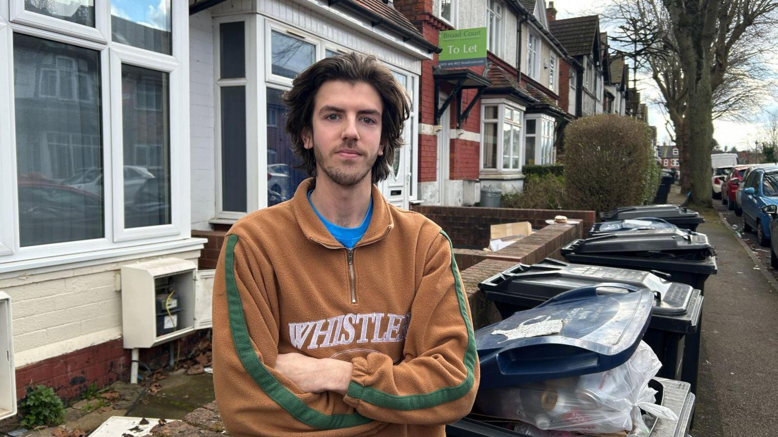 A man with a brown jacket and blue T-shirt stands with his arms crossed in front of a house. A row of bins and trees can be seen beside bin.