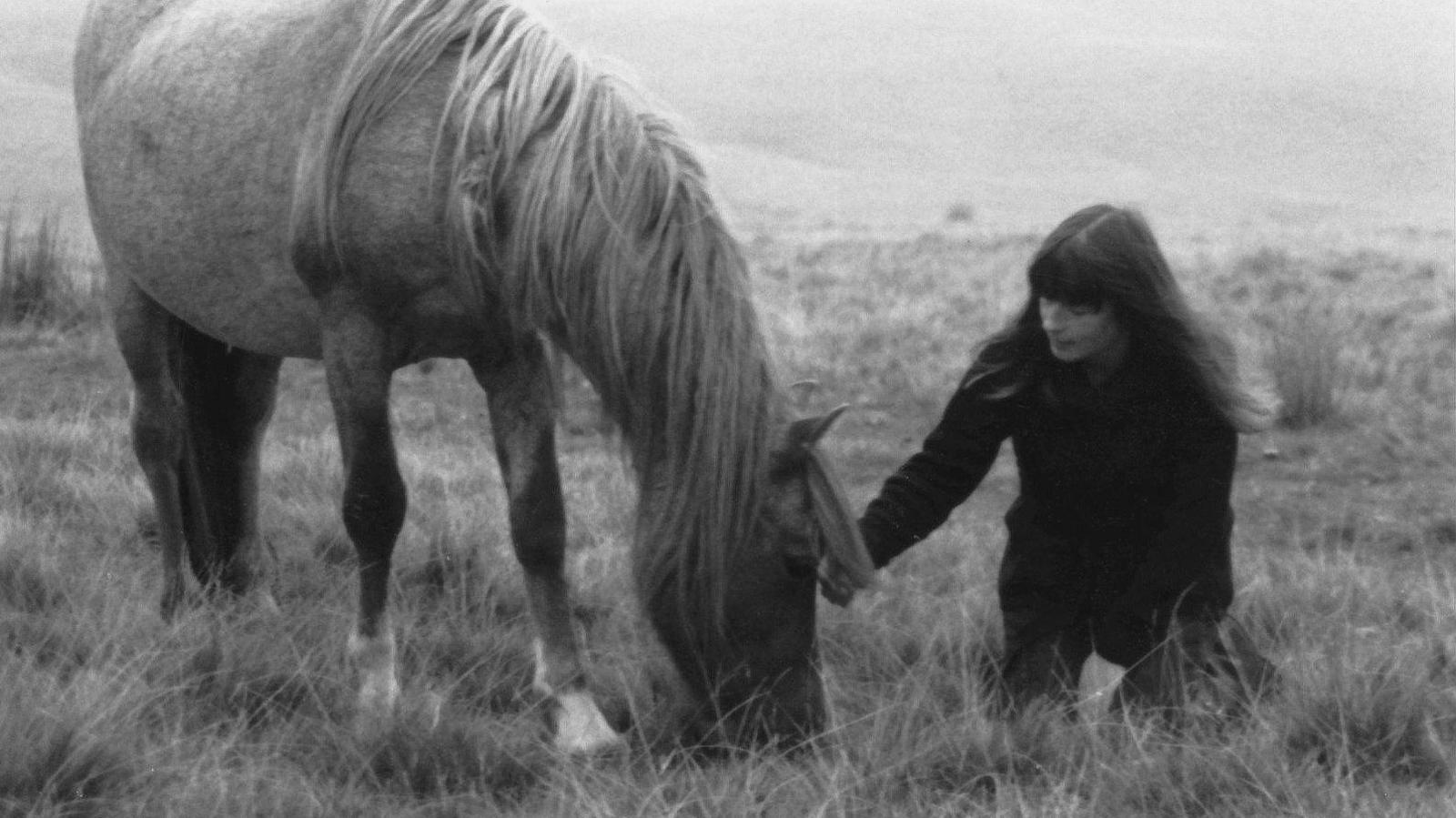 A black and white photo of Ginny and a horse on a grassy mountainside. Ginny is crouching down with her hand outstretched towards the horse, and her long hair is blowing slightly in the wind. 