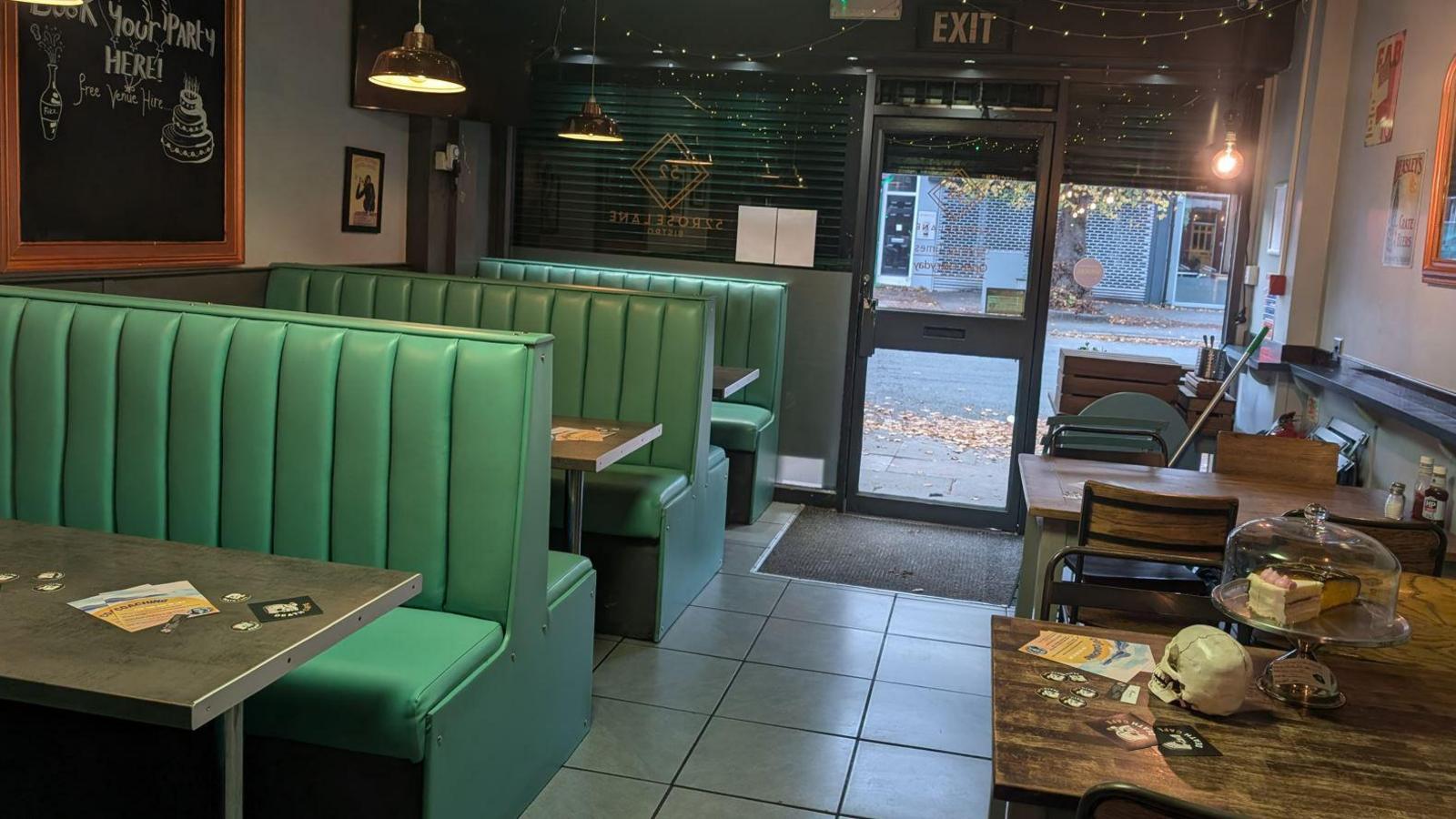 A café with green booths and mood lighting with wooden tables to the right with skulls on and cake