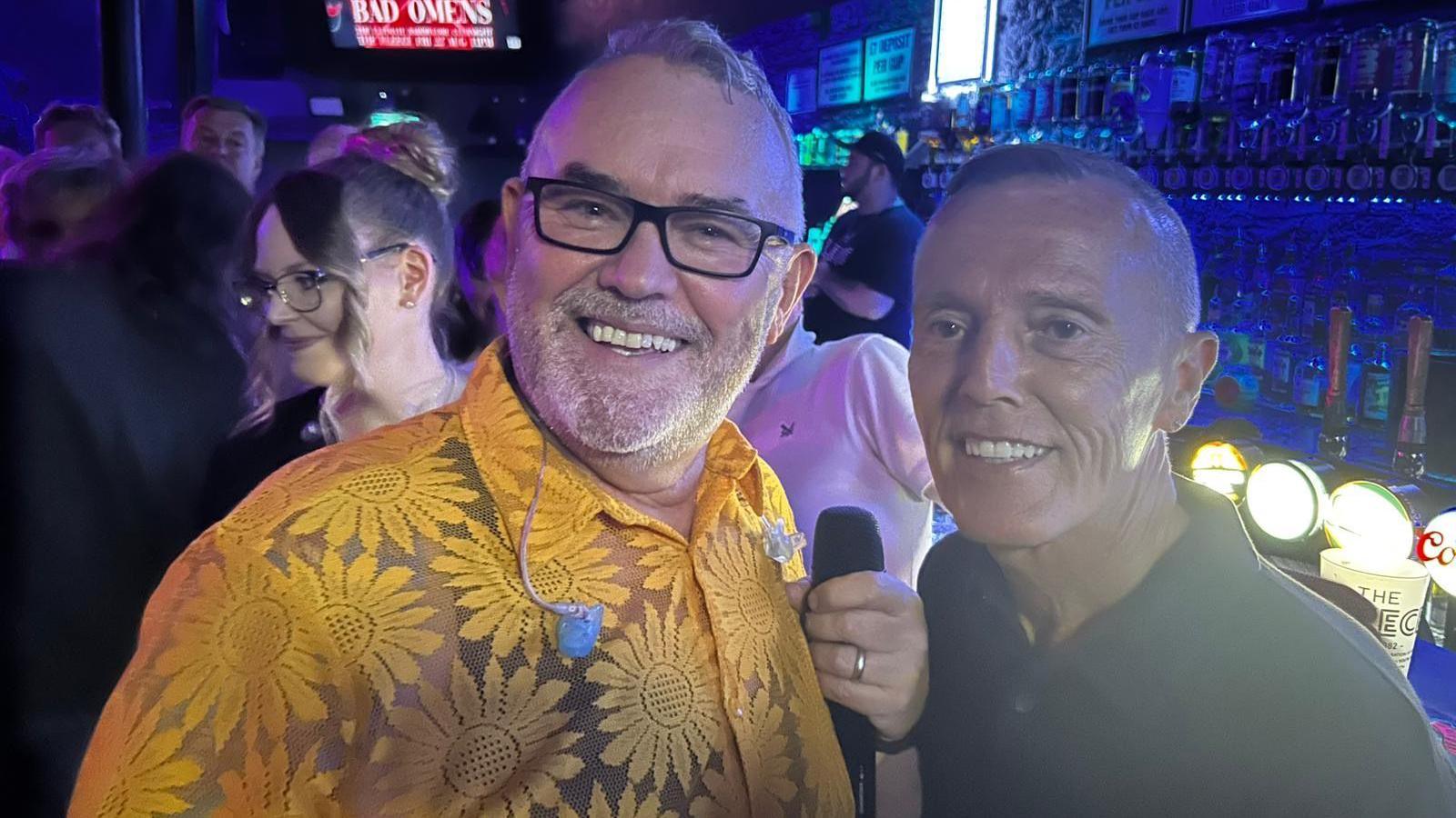 Two men stand next to each other for a photo in a bar, both smiling at the camera.