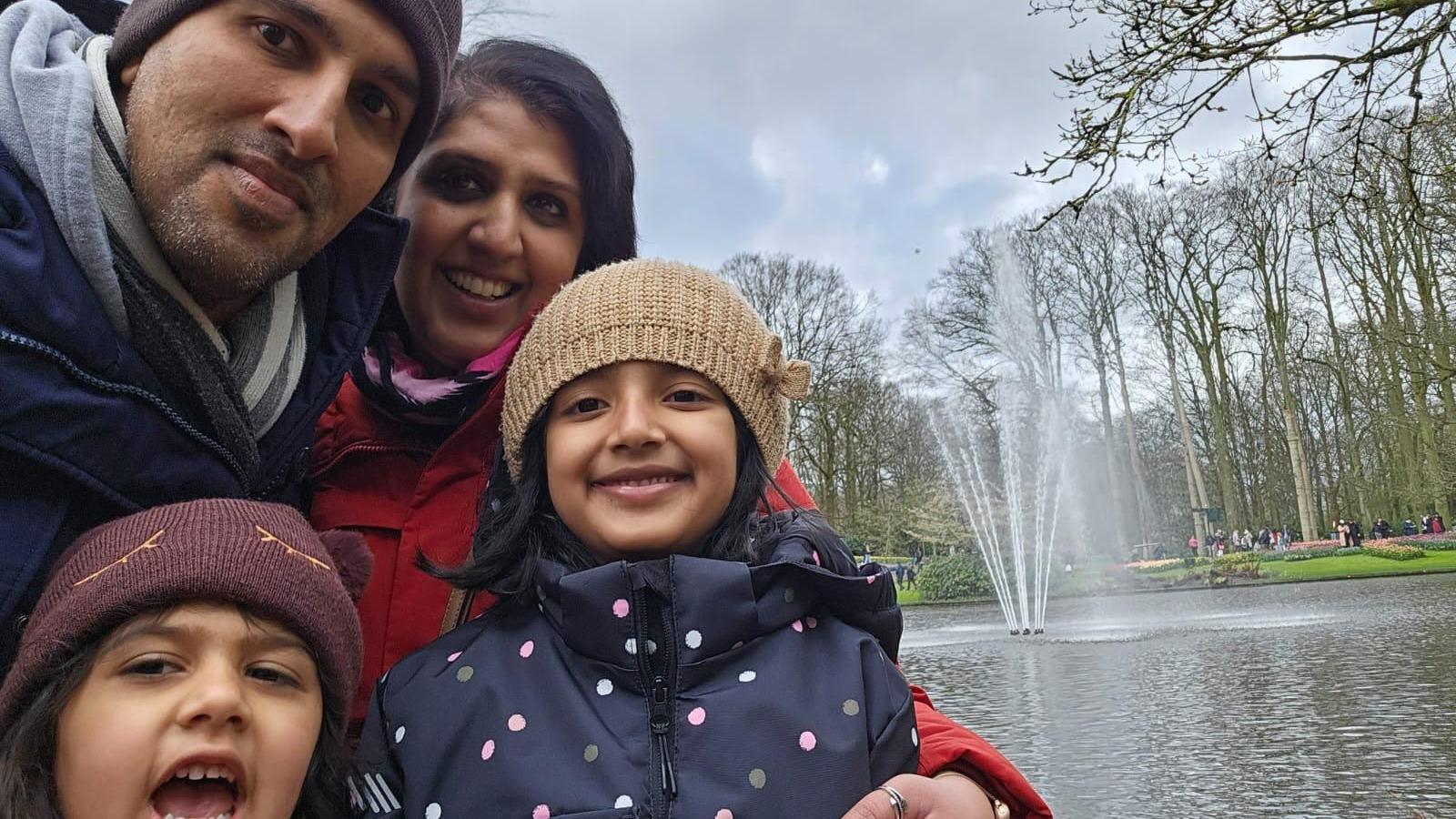 A family in a park, a man to the left, a woman, and two children at the front. They are all wearing winter coats, the children have hats on, the man has a hat on and Priyanka has her hair down and is wearing a red coat. They are all by a park with a water and a water feature in the middle. 