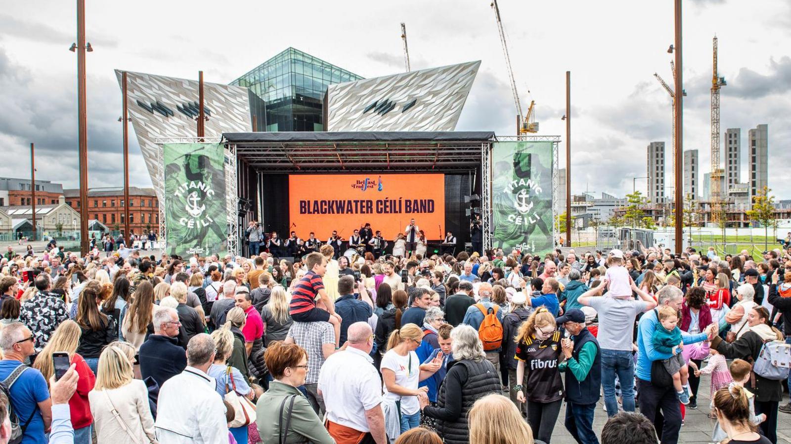 The grey and glass triangular Titanic Musuem sticks out the back of a stage and orange screen with big writing that says 'BLACKWATER CÉILÍ BAND' on it. Hundreds of people are in front of the stage, standing and some are dancing.