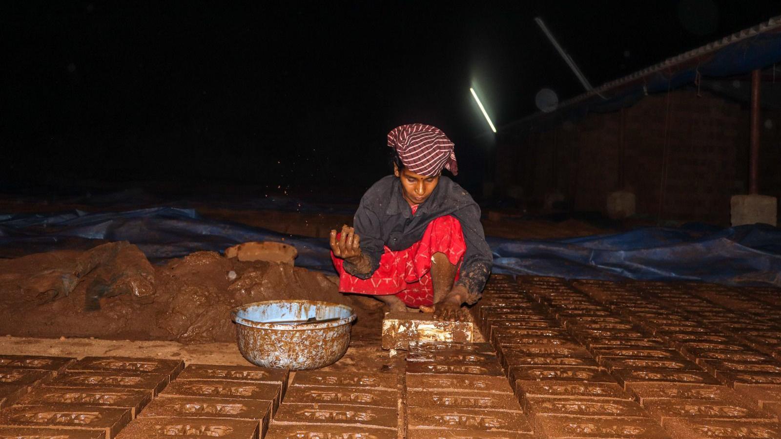 A woman wearing a red dress and grey shirt is making brown coloured bricks. There is a pile of brown mix on her side which she uses to make bricks.