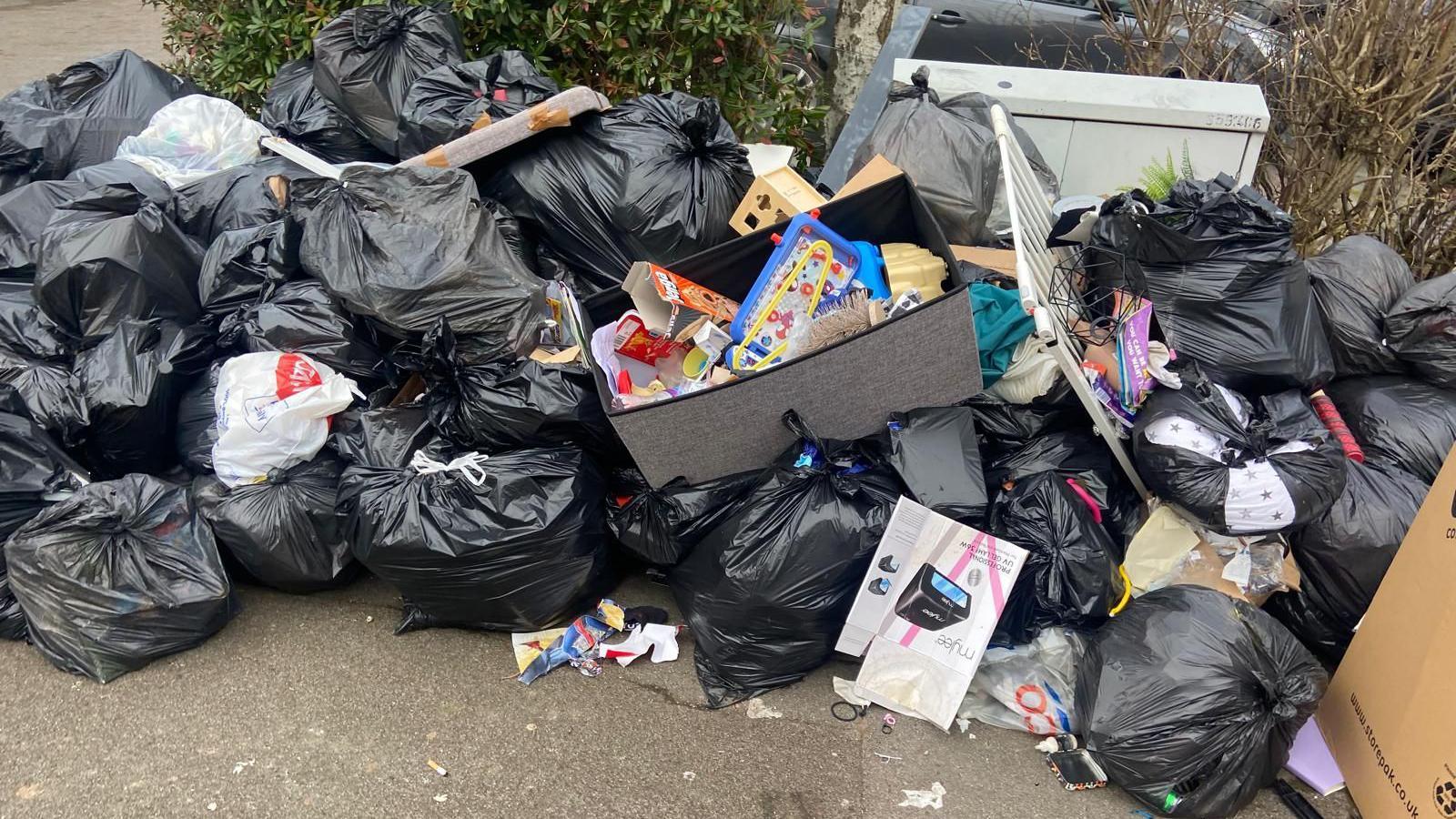 A huge pile of black binbags on a pavement. Many of the bags have split open, there is rubbish falling out. There are cardboard boxes and a white child gate on top of the bags.