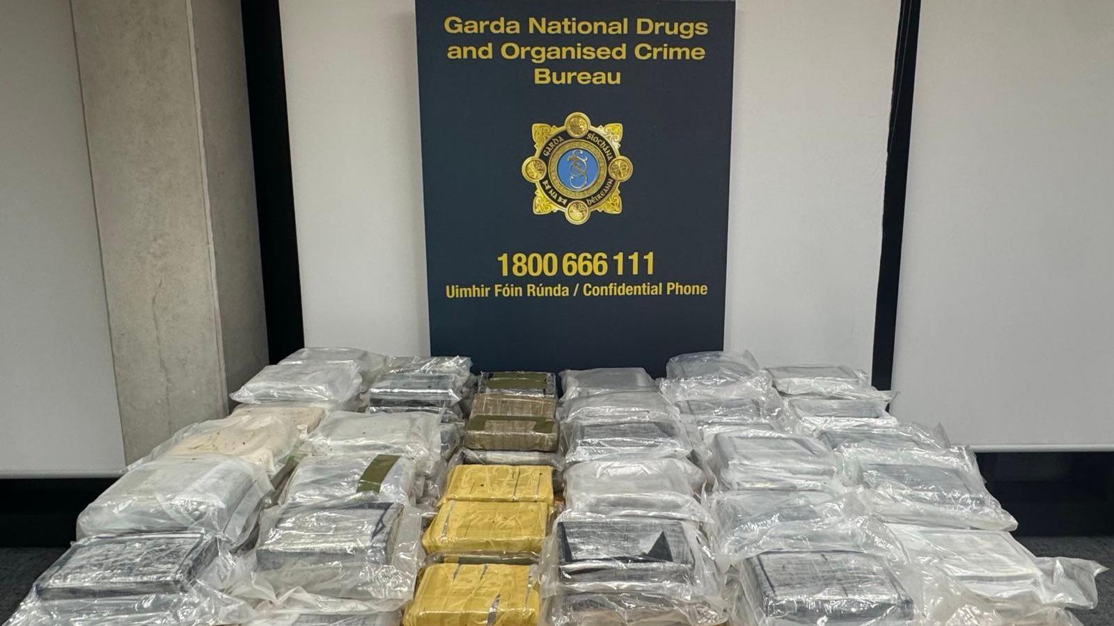 A display of suspected cocaine packets seized earlier in County Laois.  The drugs are stacked in small bundles, mostly wrapped in clear plastic but some are covered in sticky tape.  The are placed in front of a navy and yellow sign which says: "Garda National Drugs and Organised Crime Bureau"
