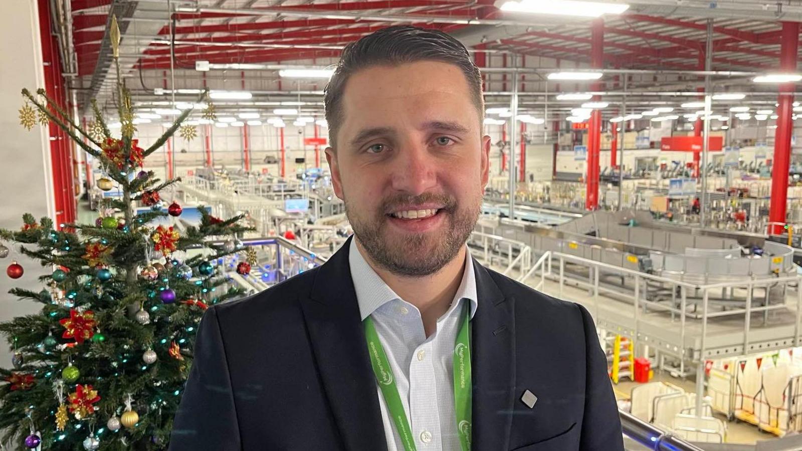 Jay Brooks pictured overlooking the Medway Mail Centre. He is stood in front of a decorated Christmas tree, and is wearing a light blue shirt and a navy suit jacket. Jay is also wearing a green lanyard.