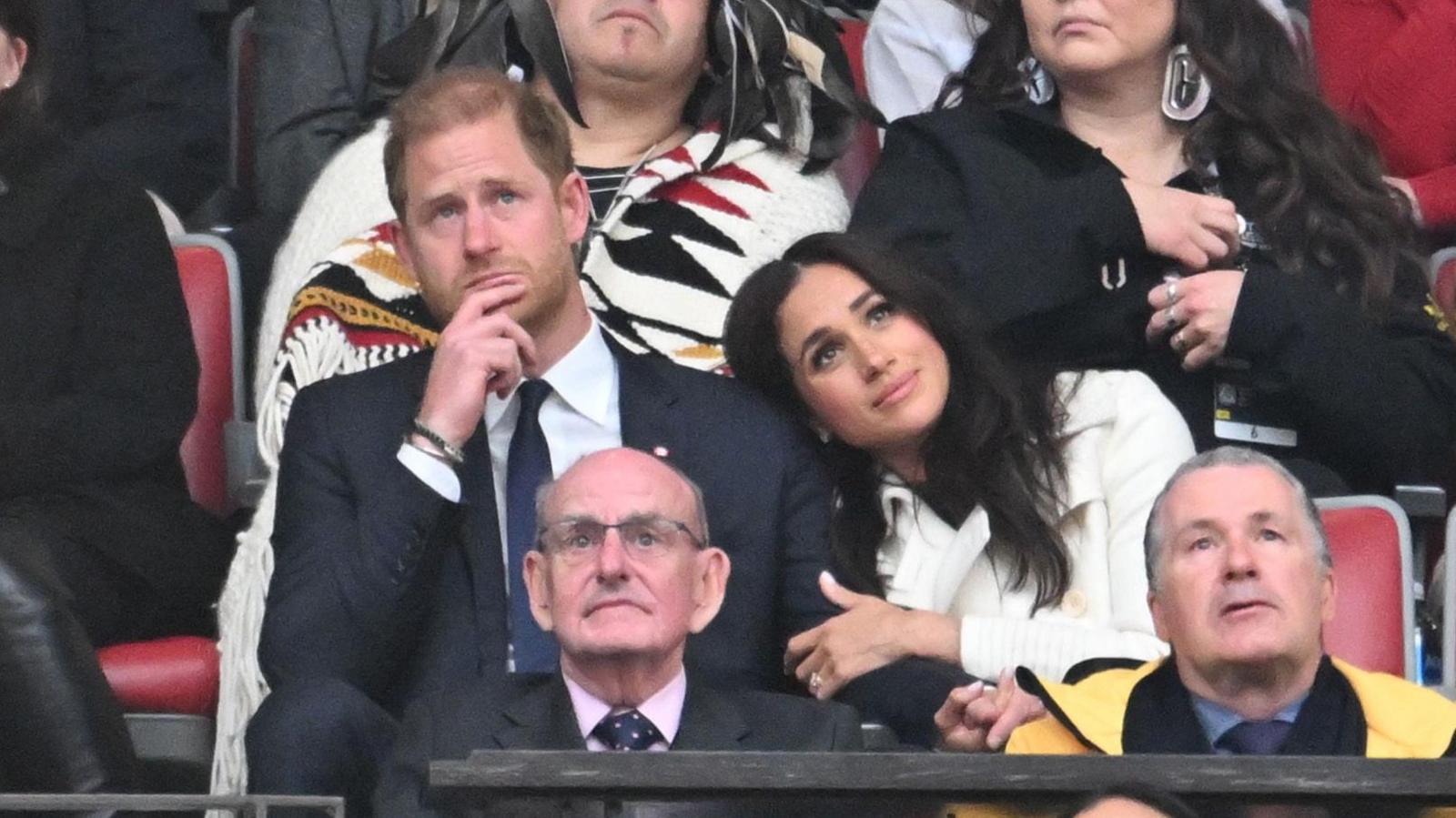 In the middle of a stadium crowd, Prince Harry, wearing a suit, looks into the distance with his finger on his chin while Meghan Markle, wearing a white coat, leans against him and clutches his arm