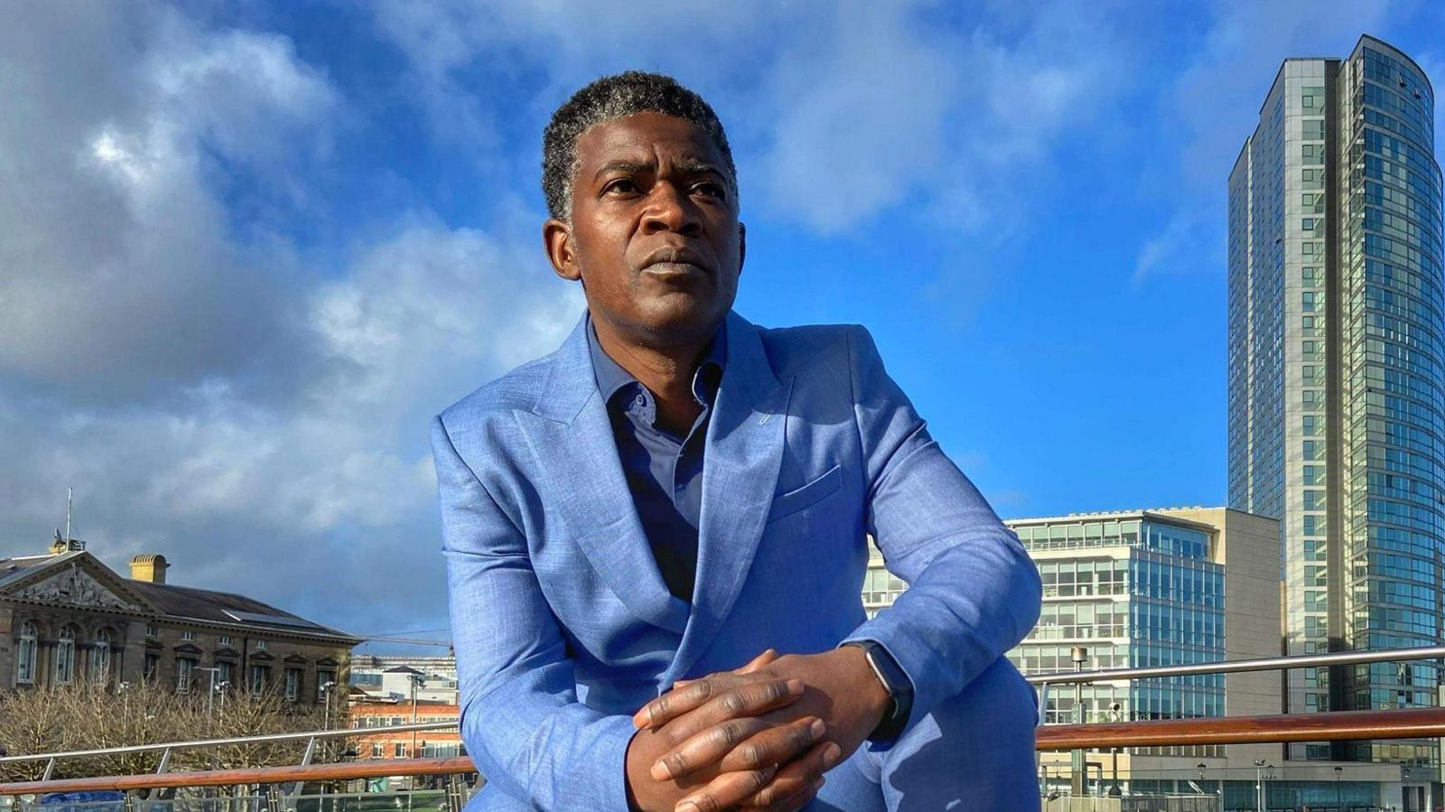 A black man pictured wearing a sky blue suit and trousers, he has his hands closed and is wearing a black apple watch. 