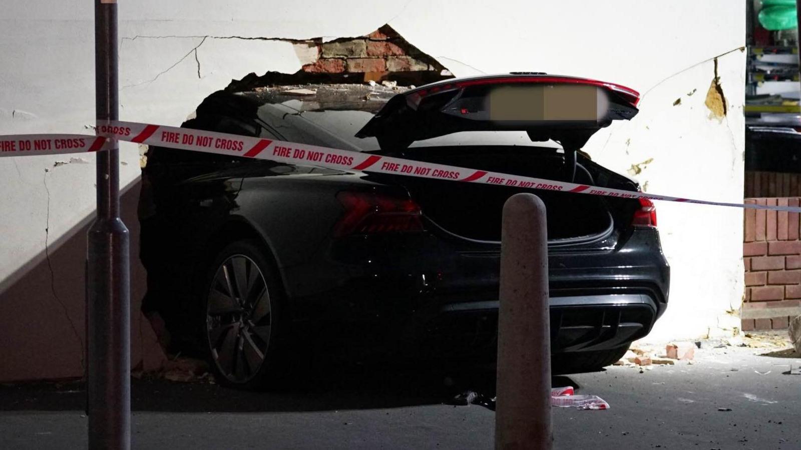 The end of a black car is visible with the rest having smashed through a white wall. 