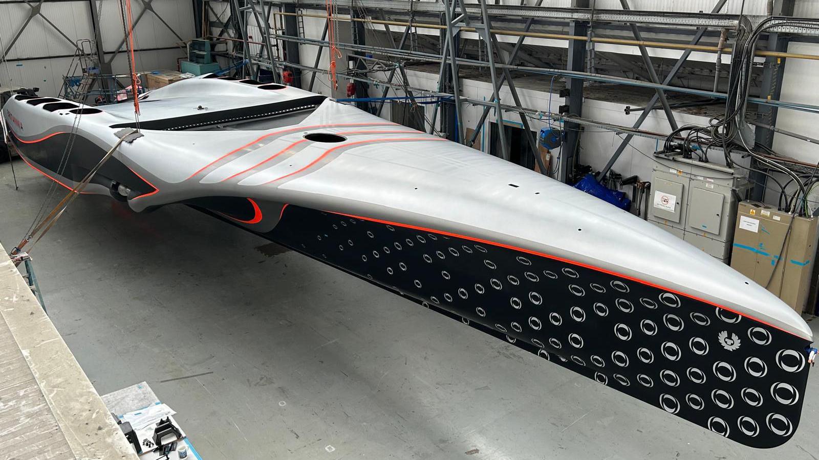 The finished hull of AC75 RB3 as seen from above. The boat is sat on stilts and is grey on it's deck and has a blue and white hull