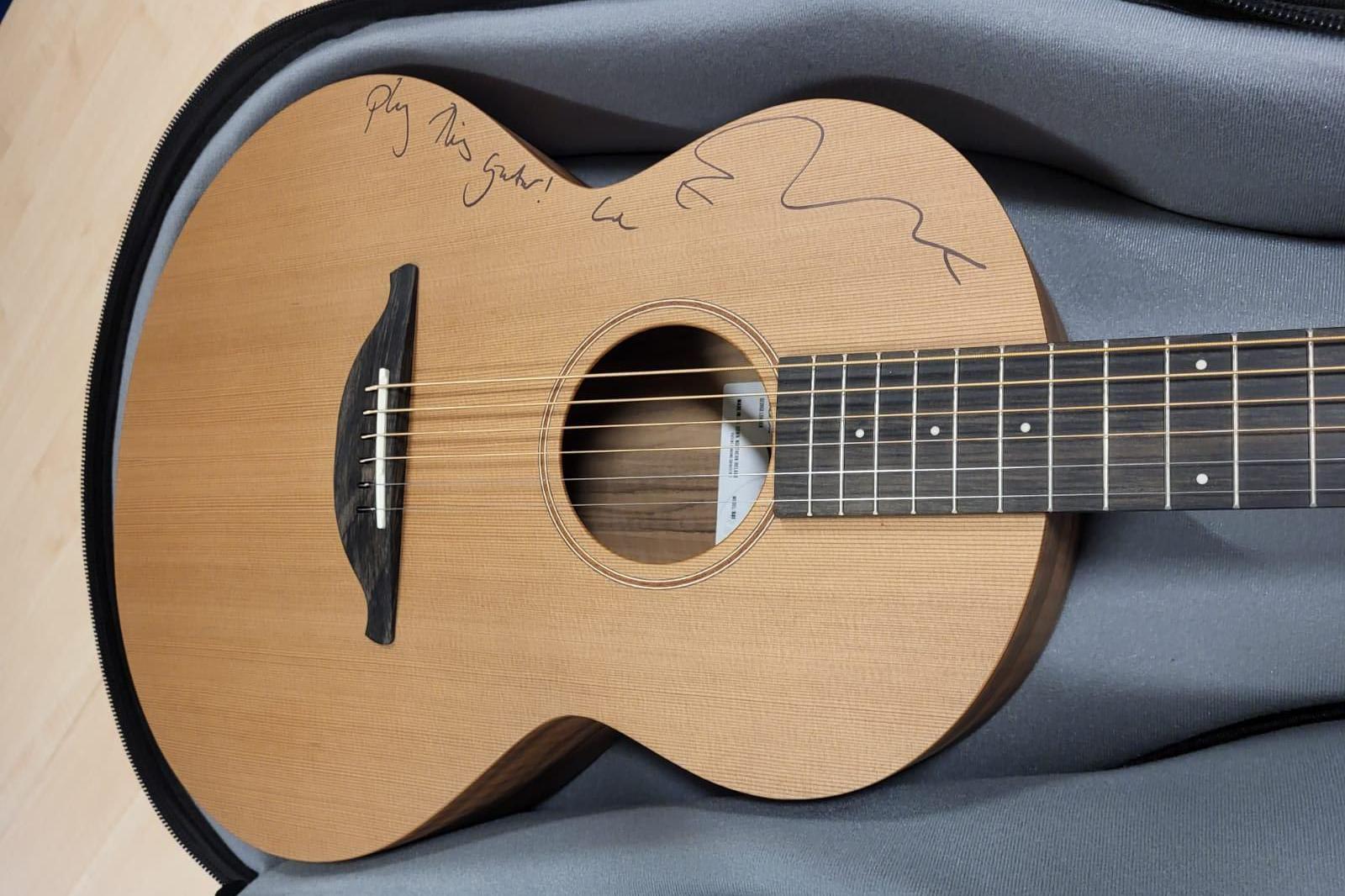 An acoustic guitar in a guitar case with the phrase Play This Guitar and Ed Sheeran's autograph written in felt tip across the top